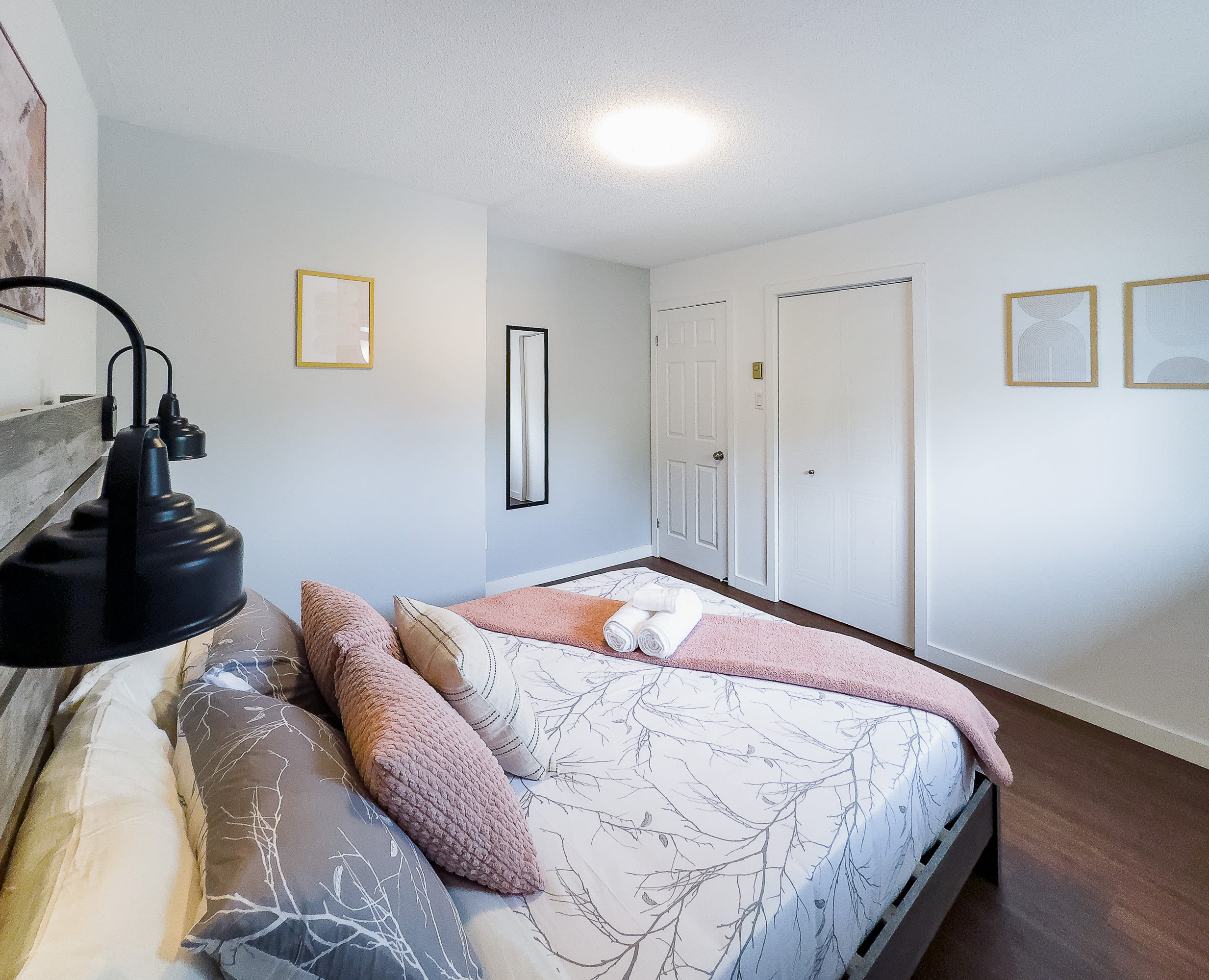 A close-up view of a rustic wooden bed with white and gray patterned bedding, accented by pink throw pillows and a pink blanket. A white door and a tall mirror are visible in the background, with dark hardwood floors.