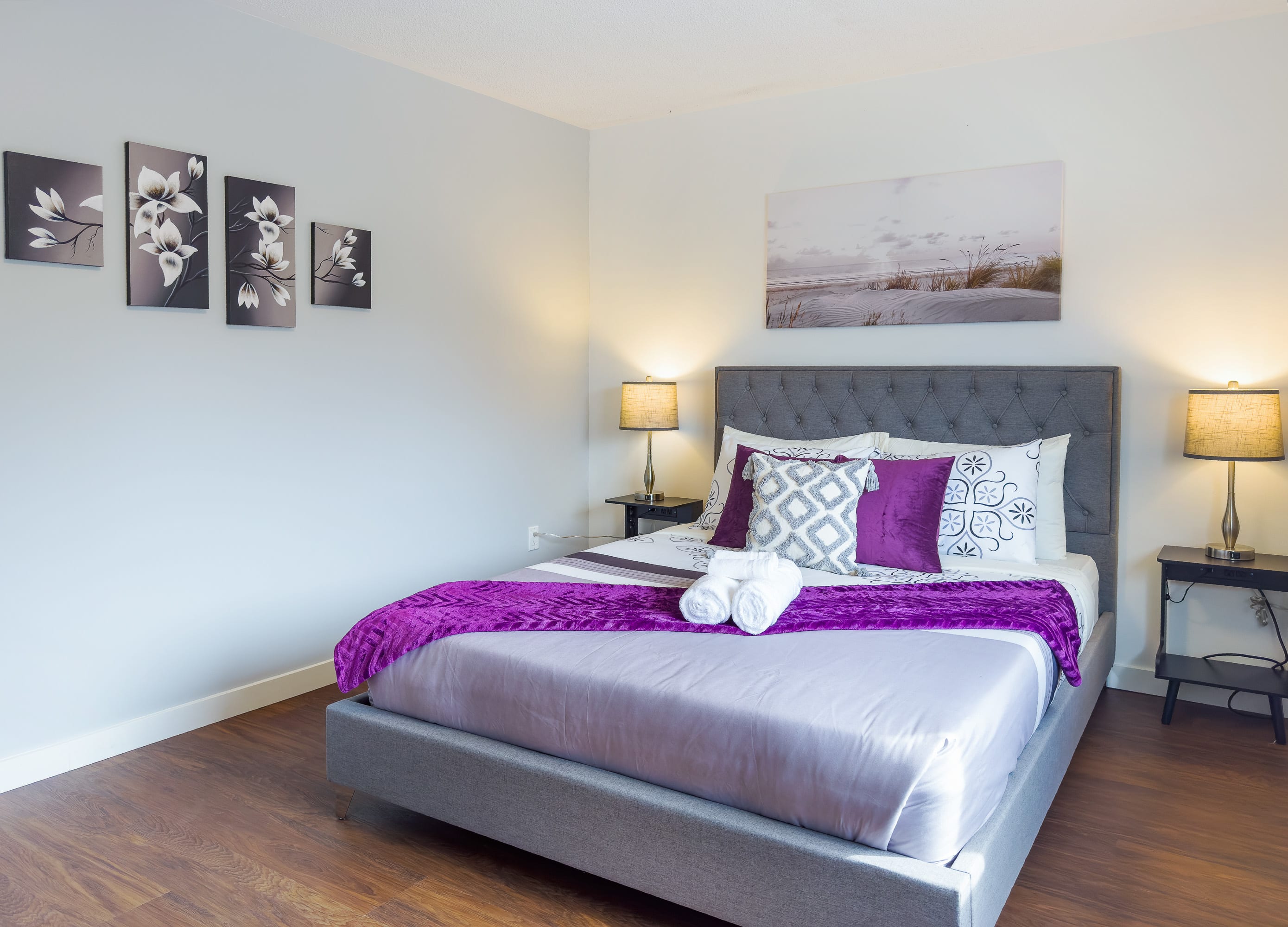 A wide shot of a bedroom featuring a gray tufted upholstered bed with purple and white bedding, flanked by two nightstands with lamps. Decorative wall art hangs above the bed and on an adjacent wall, complementing the light gray walls and dark hardwood floors.