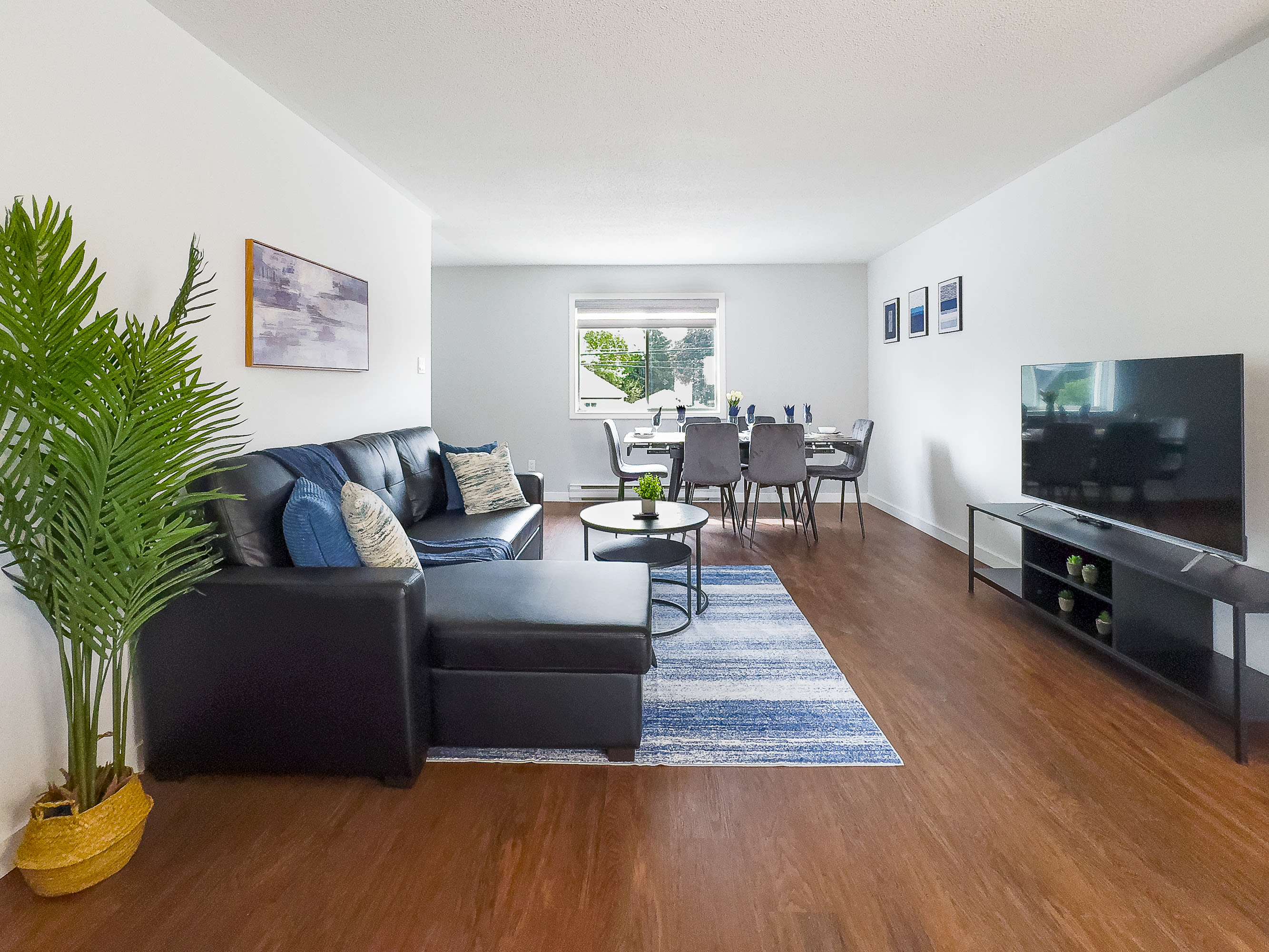 Spacious open-concept living room with a black sectional sofa, blue and white striped rug, and a large flat-screen TV on a black media stand, flowing into a dining area.