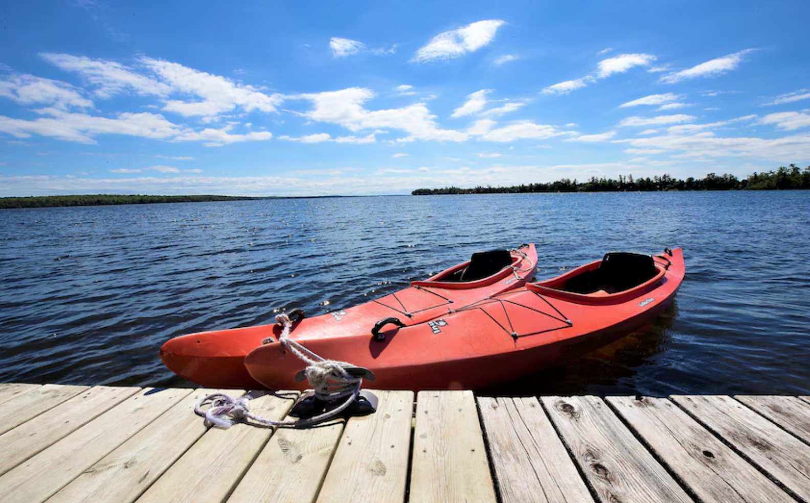 Two vibrant red kayaks are moored to a rustic wooden dock, extending into a calm, expansive lake under a bright blue sky with scattered white clouds. A distant treeline marks the far shore.