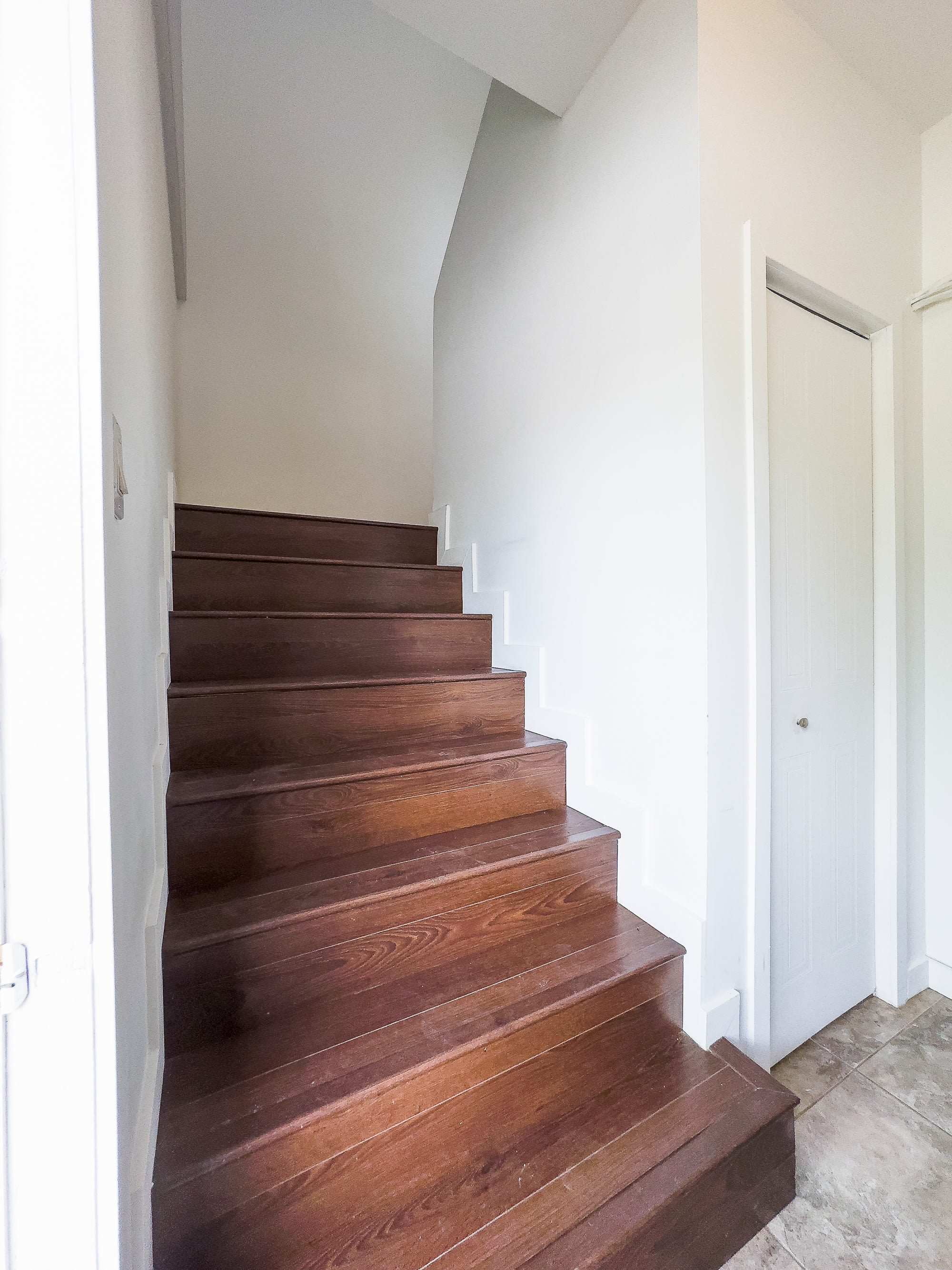 A simple wooden staircase with dark stained treads and white risers, leading upstairs. The walls are painted white, creating a clean and minimalist look.