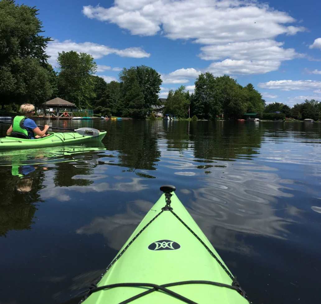 A scenic outdoor shot from the perspective of a kayak on a calm lake, with another person kayaking ahead. The background features lush green trees and houses under a bright blue sky with fluffy white clouds.