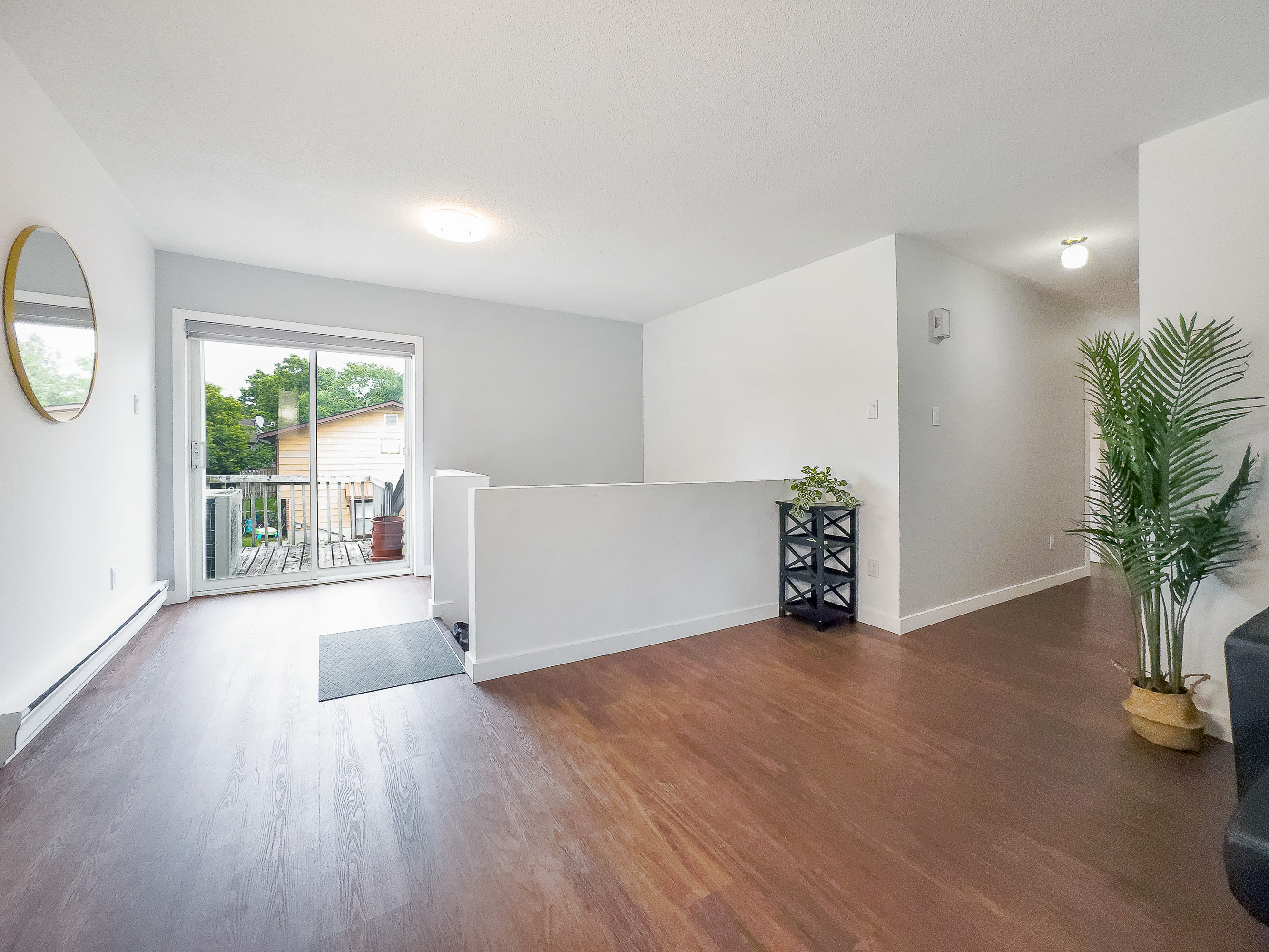A bright living area with dark hardwood floors, featuring a white half-wall separating it from an entryway. A sliding glass door leads to an outdoor balcony, and a large round mirror hangs on the wall.