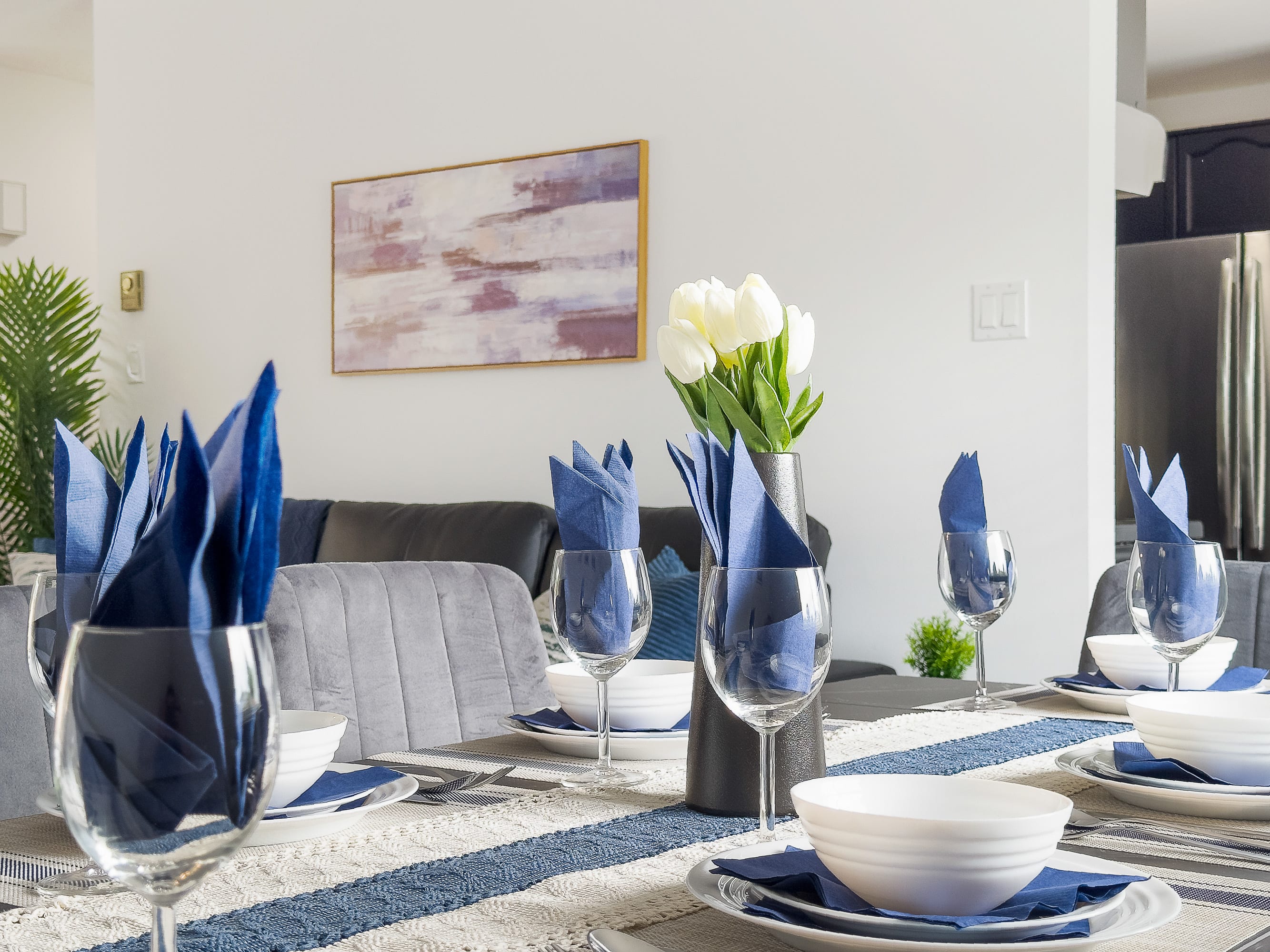 Close-up of a dining table elegantly set with white bowls, blue napkins folded into wine glasses, and a vase of white tulips, highlighting the table runner's blue stripe.