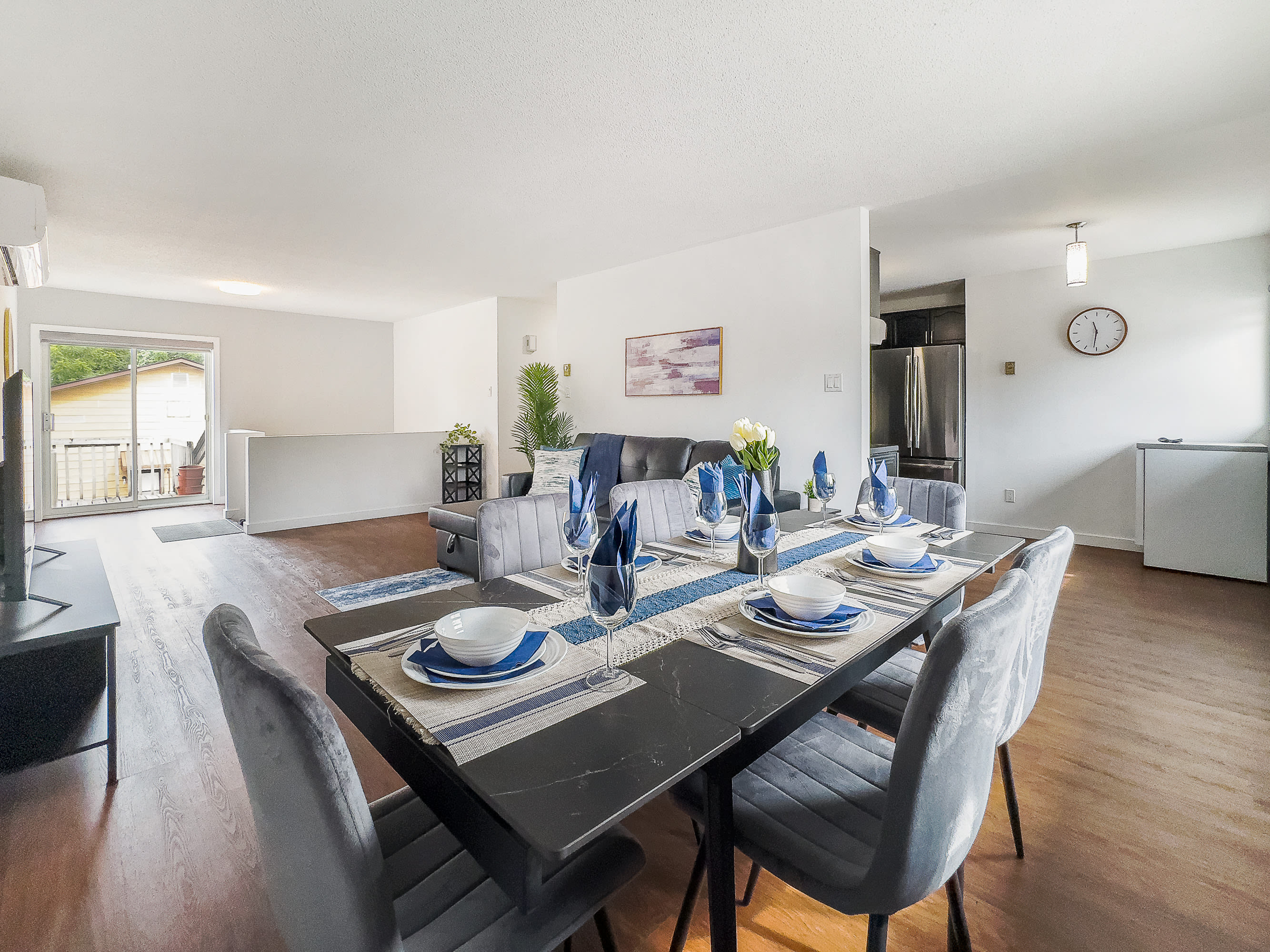 Well-appointed dining area featuring a dark rectangular table set with blue and white place settings, gray upholstered chairs, and a view into the adjacent living room and kitchen.