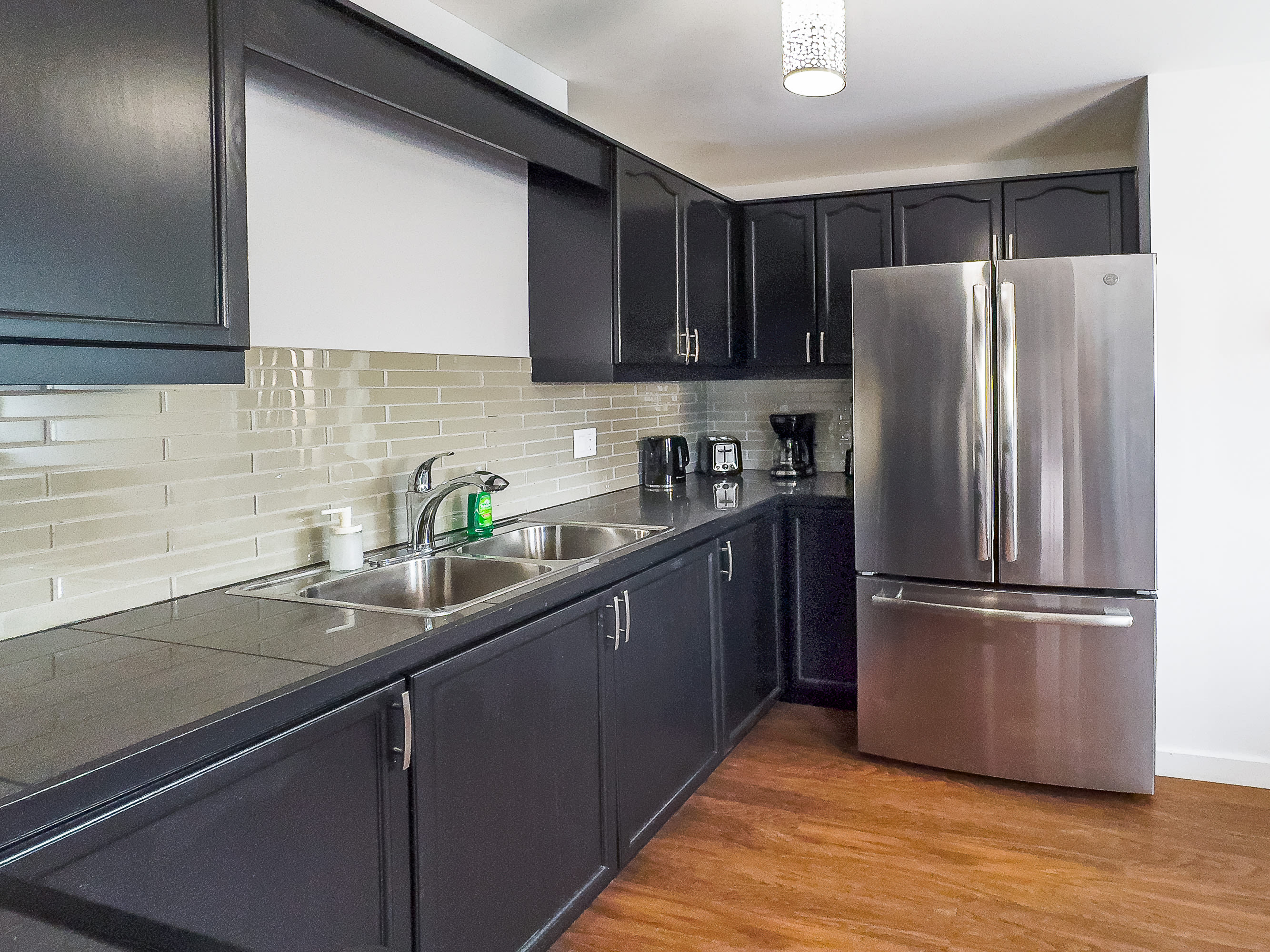 Modern kitchen showcasing dark cabinetry, black countertops, a stainless steel French door refrigerator, and a light-colored subway tile backsplash, with a double sink visible.