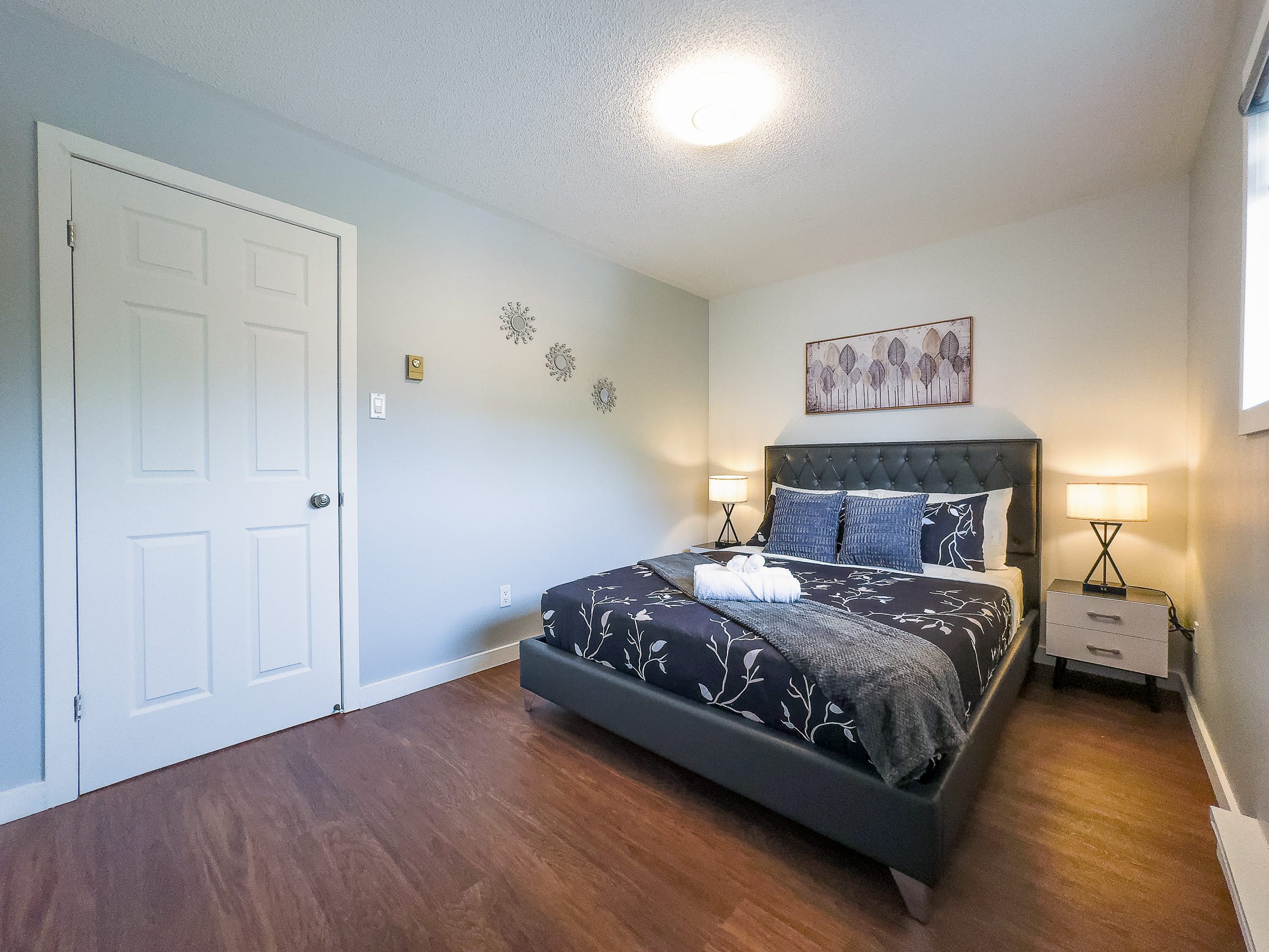 A cozy bedroom featuring a dark upholstered bed with patterned bedding, two bedside tables with lamps, and light gray walls. Dark hardwood floors complete the space, with a white door visible on the left.