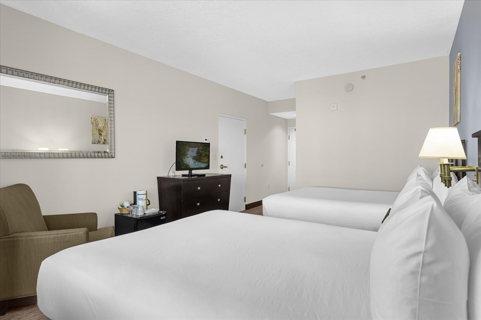 A wide view of a studio bedroom with two beds, light beige walls, and a dark wood dresser with a TV. An armchair and a mirror are visible on the left, while a door leads to another room or closet.