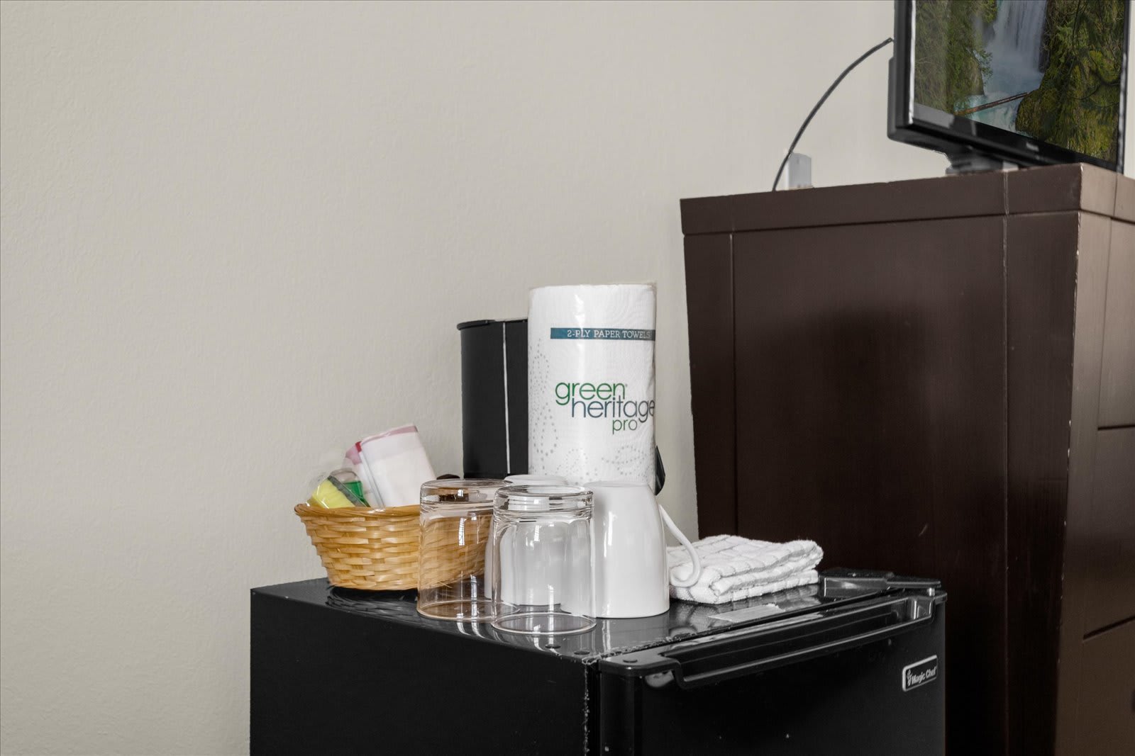 A close-up of a mini-fridge and coffee station in a bedroom, featuring a basket with glasses, paper towels, and a coffee maker. A flat-screen TV is visible on a dark wood dresser in the background.