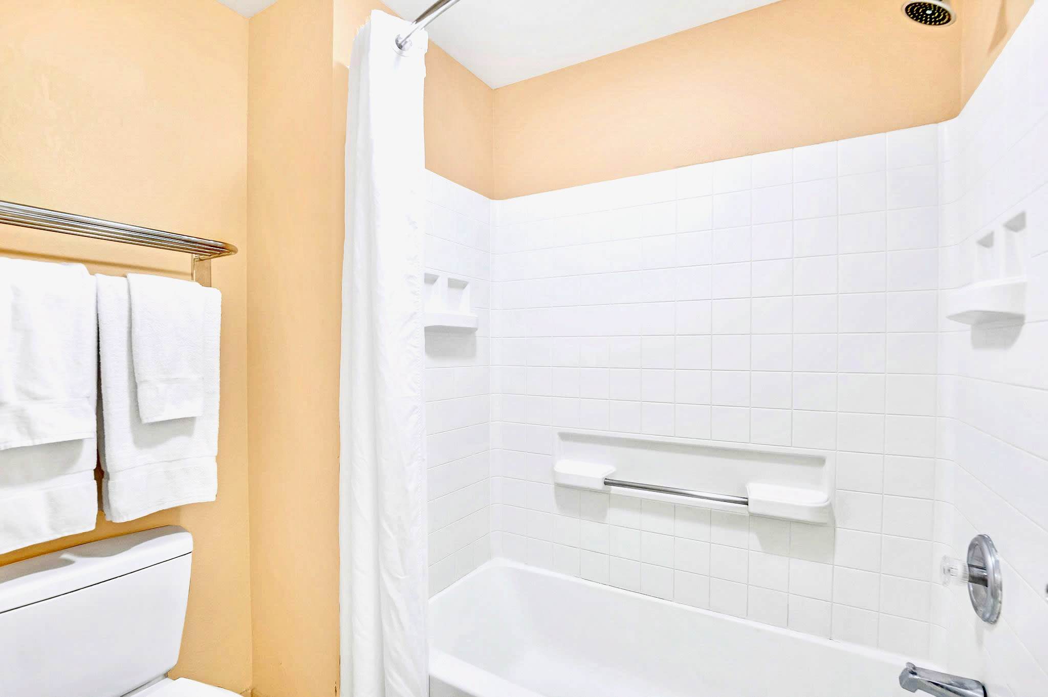 A close-up view of a clean shower-tub combination with white subway tiles and a white shower curtain, featuring a chrome shower head and grab bar.