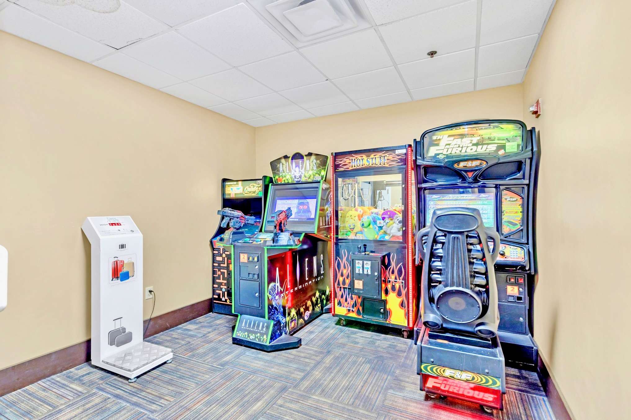 An indoor arcade room is filled with various video game machines, including racing games and claw machines, set on a patterned carpeted floor. The walls are light beige, and the ceiling has standard acoustic tiles with fluorescent lighting.
