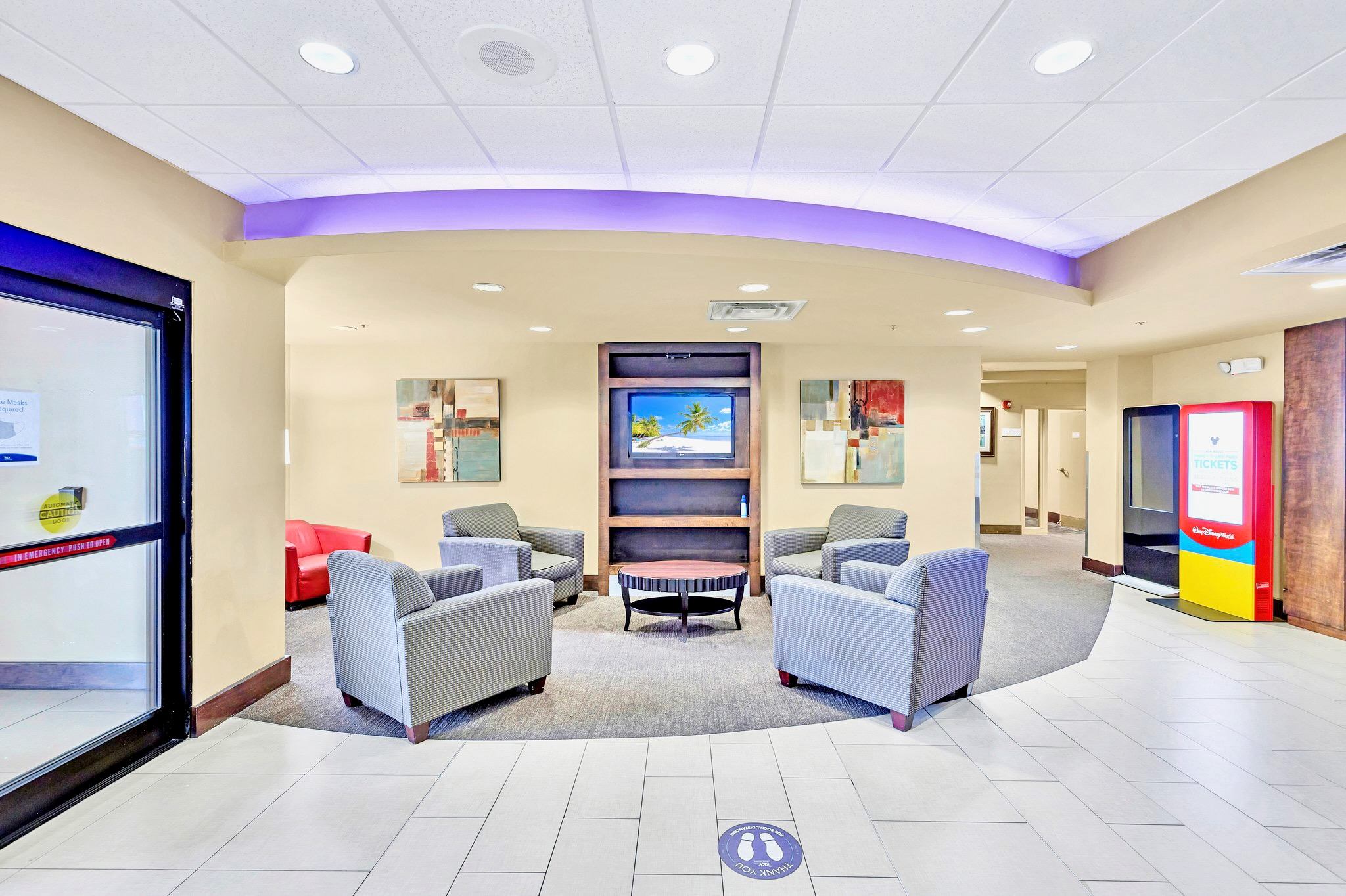 A comfortable communal lounge area features several armchairs arranged around a round coffee table, facing a wall-mounted TV within a built-in shelf. The space has a patterned carpet, light beige walls, and a curved ceiling with accent lighting.