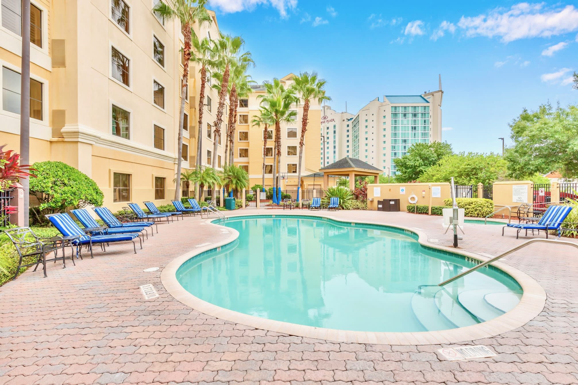 A large, freeform swimming pool with light blue water is surrounded by a brick paver deck, several blue and white striped lounge chairs, and tall palm trees. A multi-story building with light yellow walls and numerous windows stands in the background under a bright blue sky.