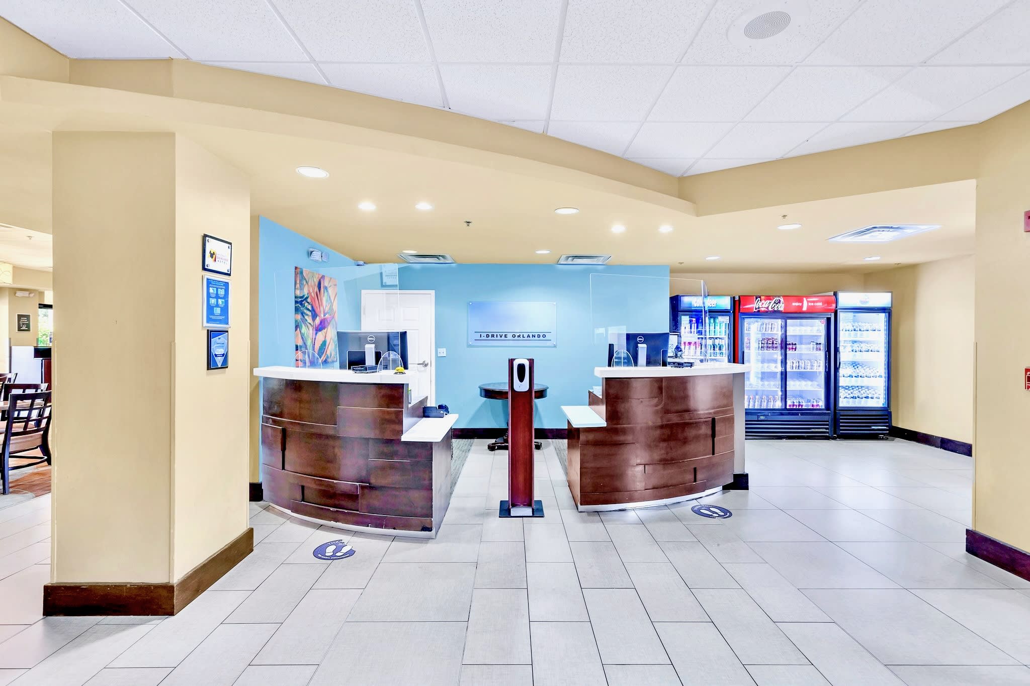 A bright reception area features two curved wooden front desks with computers, separated by a hand sanitizer station. The floor is tiled, and large refrigerators stocked with beverages are visible against a light blue accent wall.