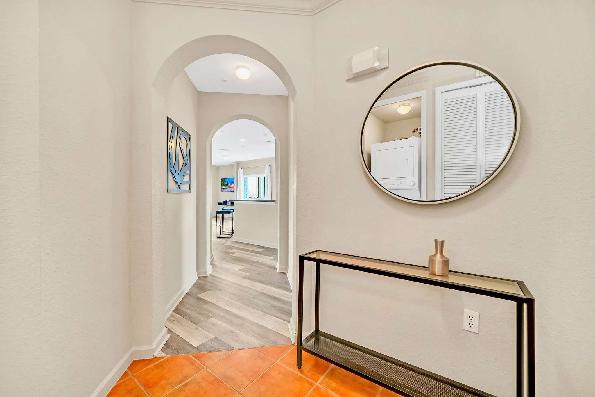 Inviting foyer with terracotta-colored floor tiles transitioning to wood-look flooring through an arched doorway. A black console table with a gold vase and a round mirror adorn the wall.