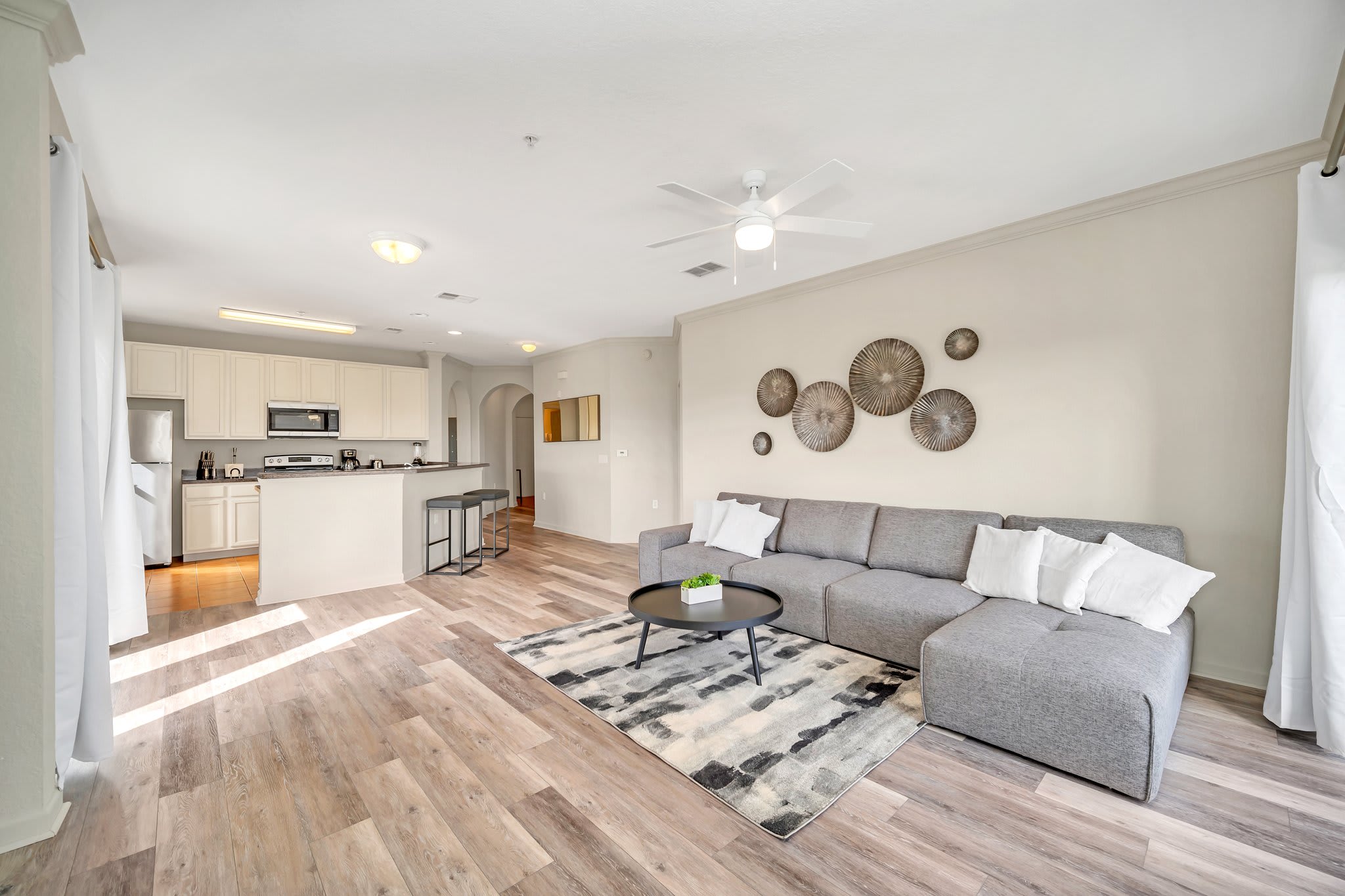 Wide shot of the open-concept living room, showcasing the grey sectional sofa, coffee table, and decorative wall art. The kitchen with its breakfast bar is visible on the left, and a hallway archway is on the right.