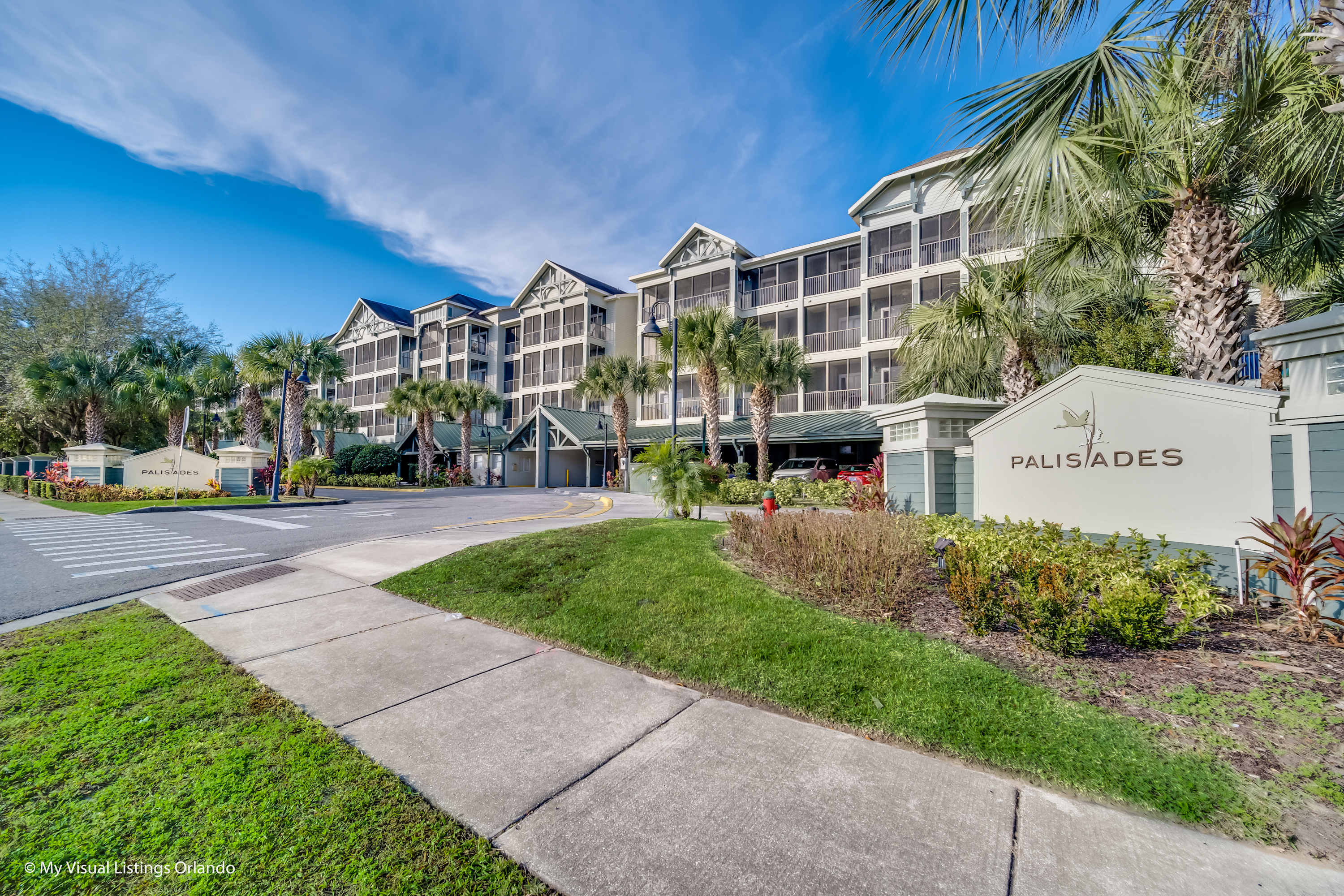 The front exterior of a multi-story condominium building with light green siding and screened balconies, surrounded by lush green grass and tall palm trees under a bright blue sky. A "PALISADES" sign is prominently displayed at the entrance.