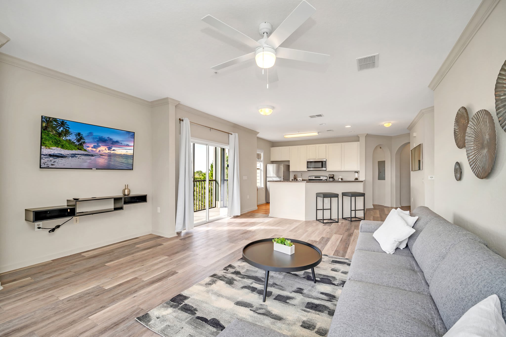 Open-concept living area featuring a large grey sectional sofa, a round black coffee table on a patterned rug, and a wall-mounted TV. The space flows into the kitchen area, visible in the background, and has access to a balcony.