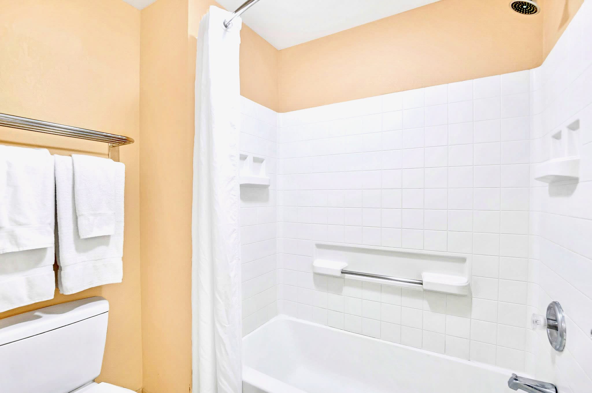 A close-up view of the bathtub and shower area in the bathroom, featuring classic white subway tiles on the walls and a clean white shower curtain. A grab bar and built-in soap dishes are installed for convenience.