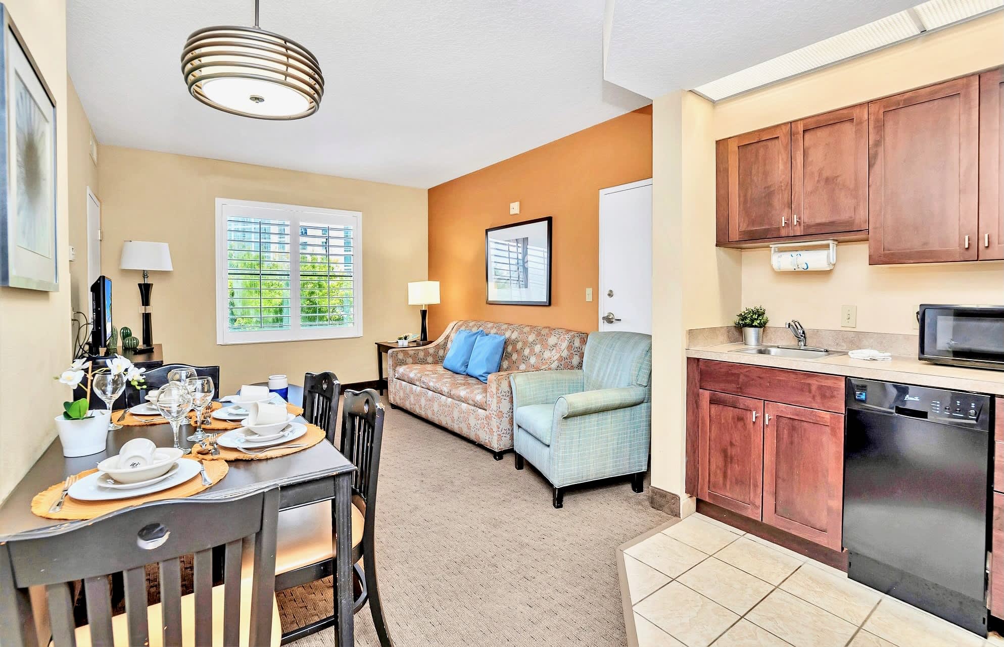 A spacious open-concept area combining a dining space, living room, and a compact kitchen. The dining table is set for four, leading to a living area with a floral sofa and armchair, while the kitchen features dark wood cabinets and essential appliances.