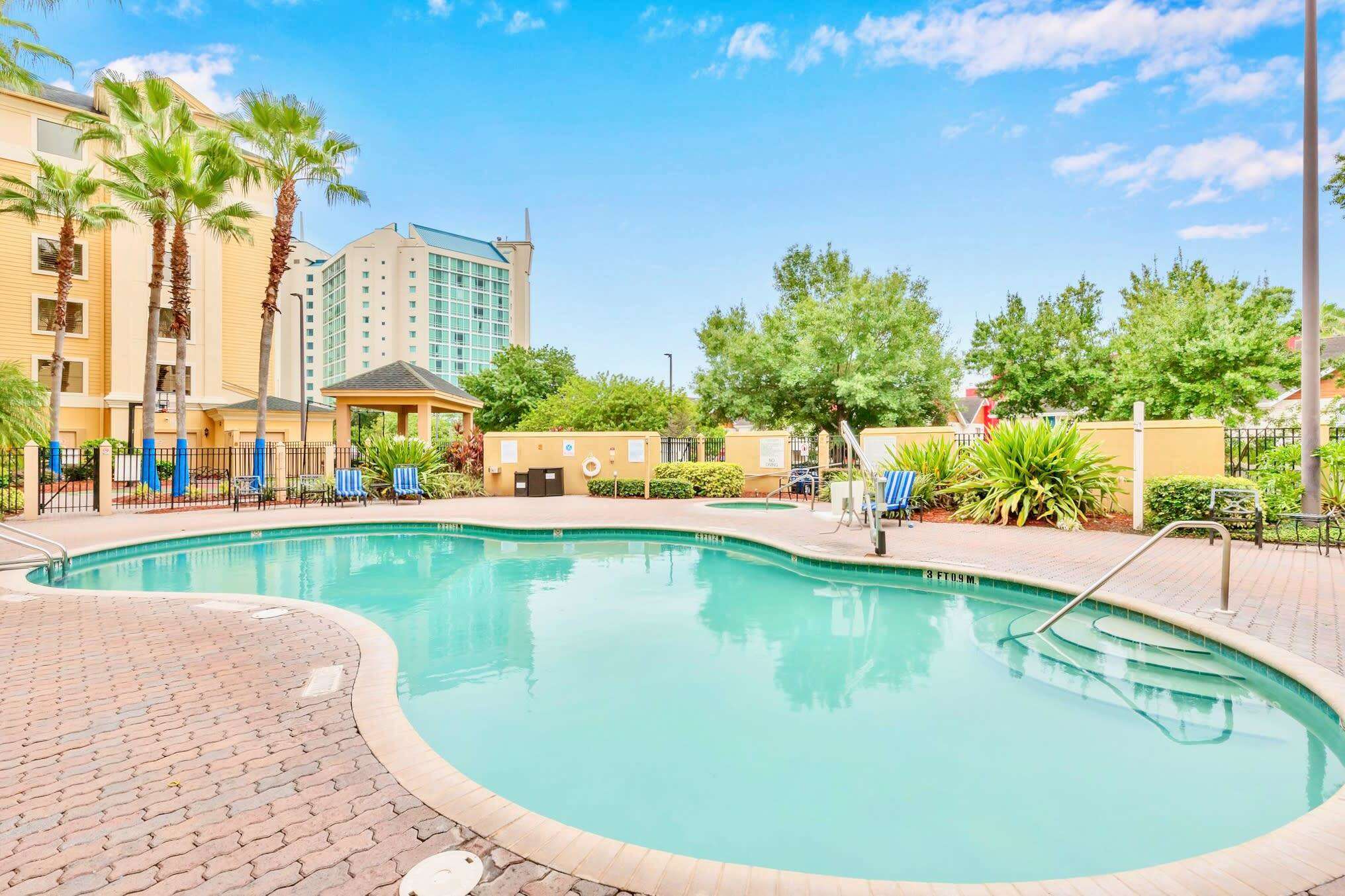 Wide shot of a large, kidney-shaped outdoor swimming pool with a hot tub visible in the background, surrounded by a brick paver deck. Palm trees and lush greenery frame the area, with a tall building under a bright blue sky.