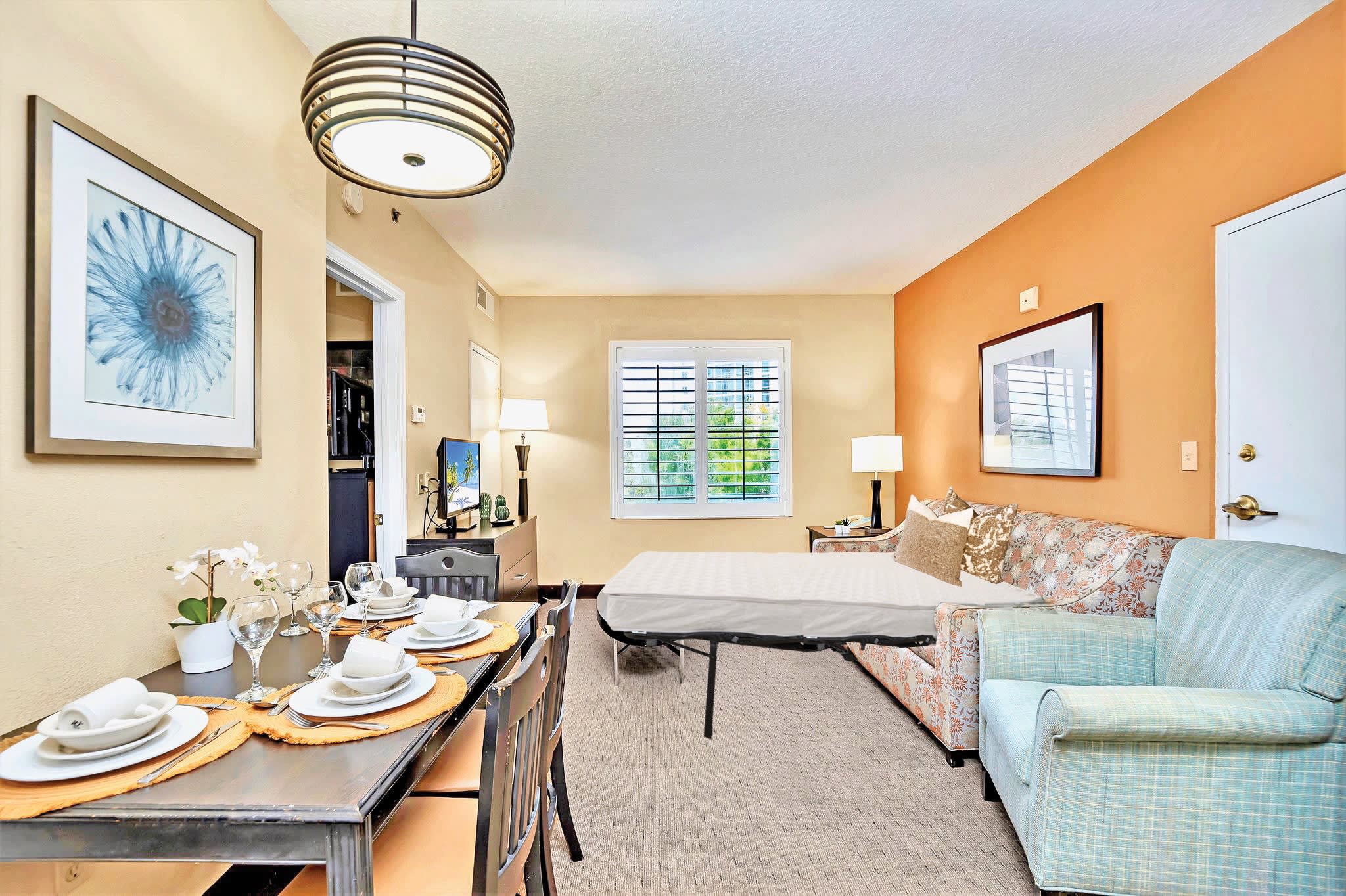 The living room area with the floral sofa converted into a pull-out bed, showcasing a mattress ready for use. The dining table remains visible on the left, maintaining the room's two-tone orange and beige walls and carpeted floor.