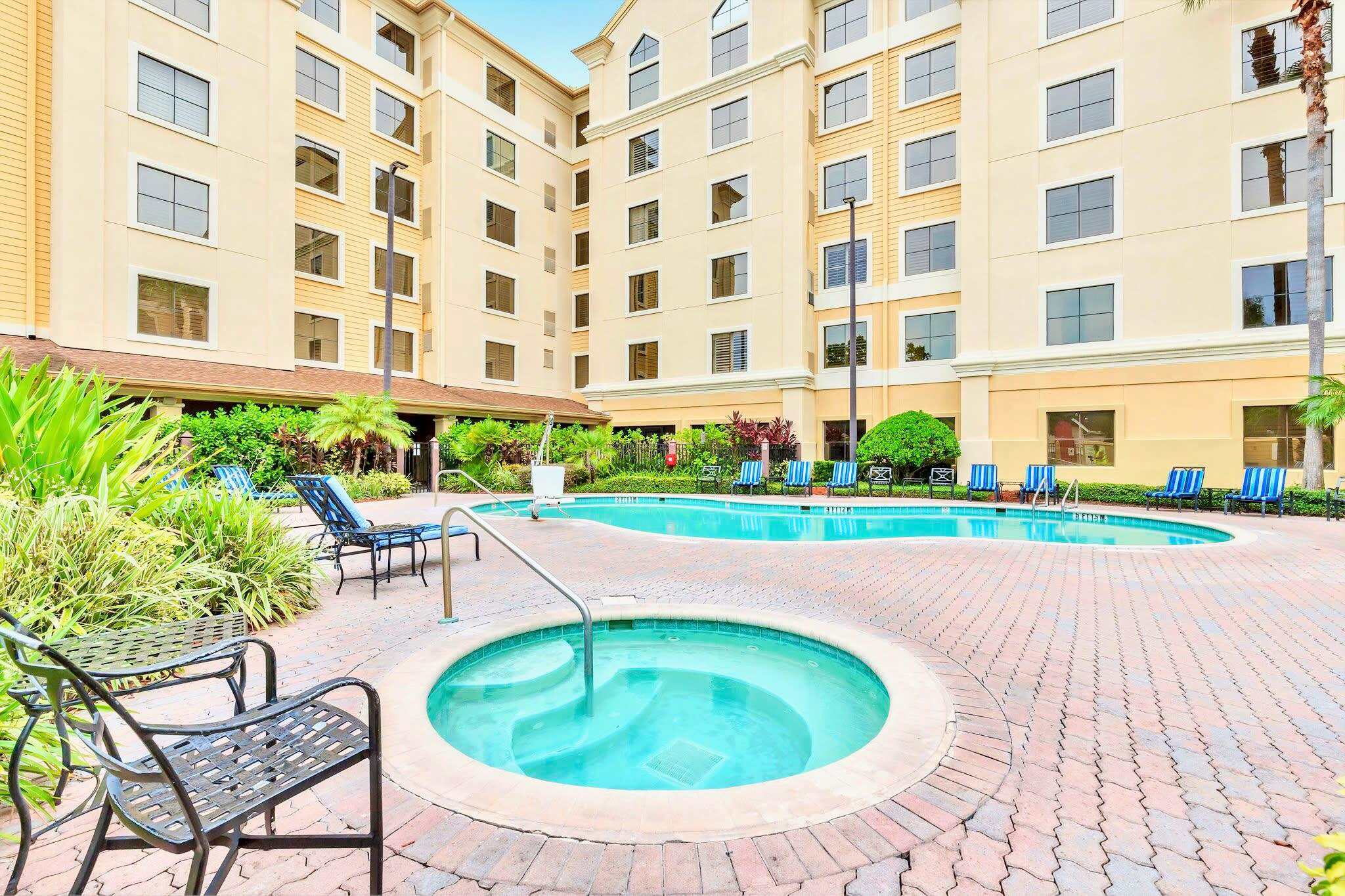 Inviting outdoor pool area featuring a large, irregularly shaped swimming pool and a circular hot tub, surrounded by a brick paver deck. Several lounge chairs are placed around the pool, with lush tropical landscaping and a multi-story building in the background.