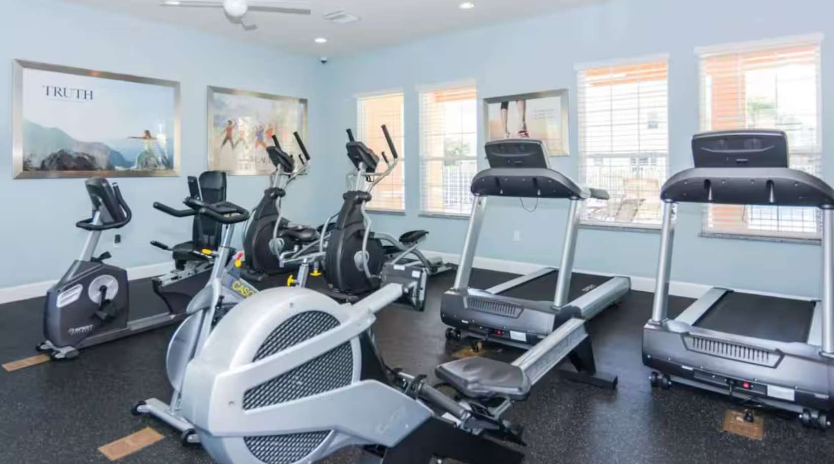 A well-equipped gym featuring multiple treadmills, elliptical machines, and stationary bikes, set on a black rubber floor with light blue walls and large windows providing natural light.