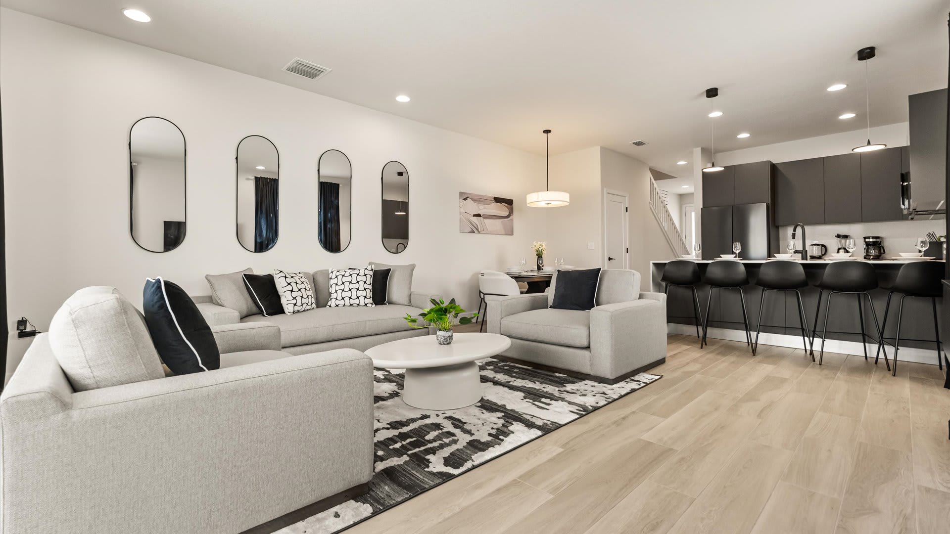 Spacious open-concept living room with light gray sofas, a white coffee table, and a black and white rug, leading to a modern dark gray kitchen with a white island and bar stools.