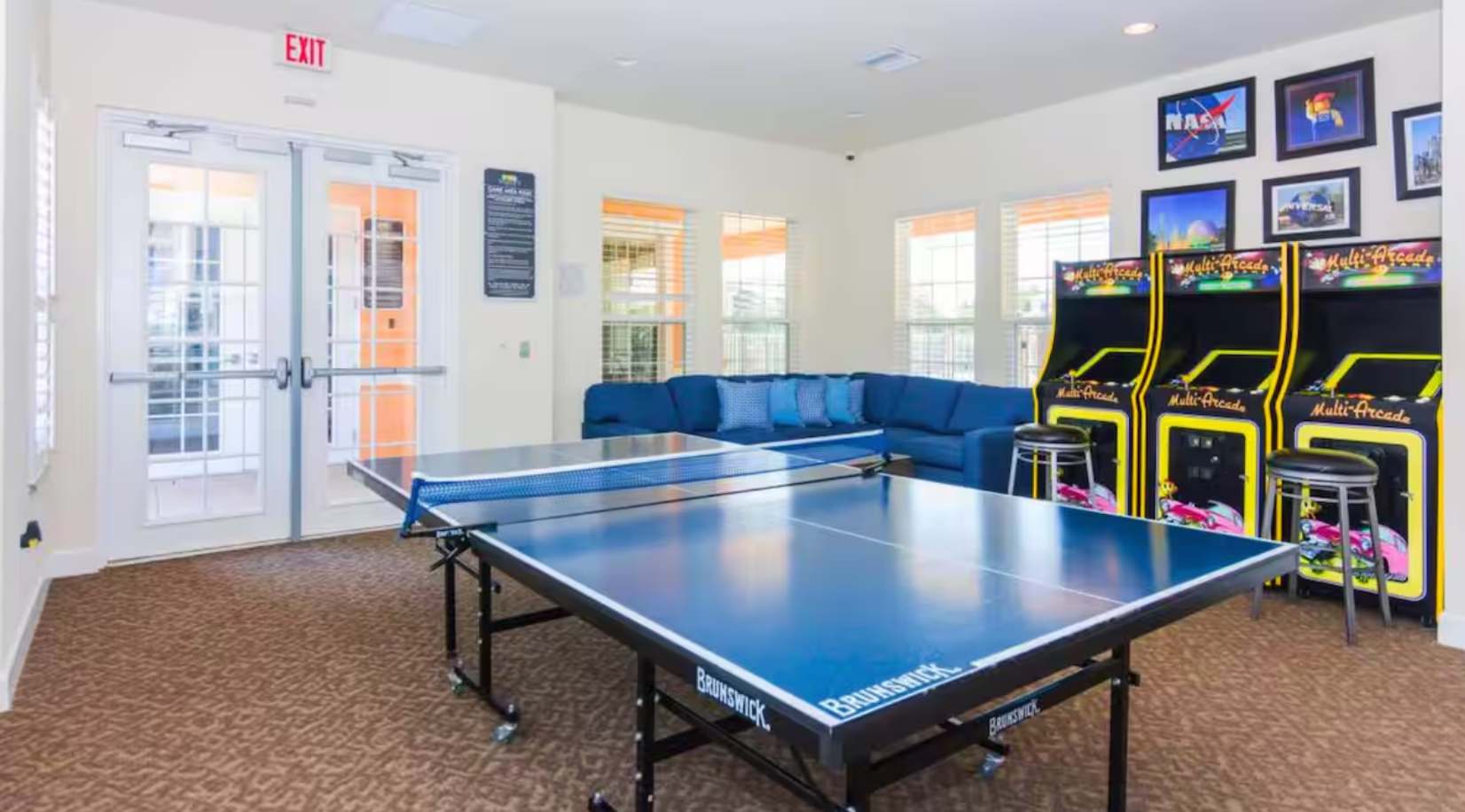 A bright game room with a blue ping pong table, a comfortable blue sectional sofa, and multiple arcade machines, featuring large windows and double glass doors leading to an outdoor area.