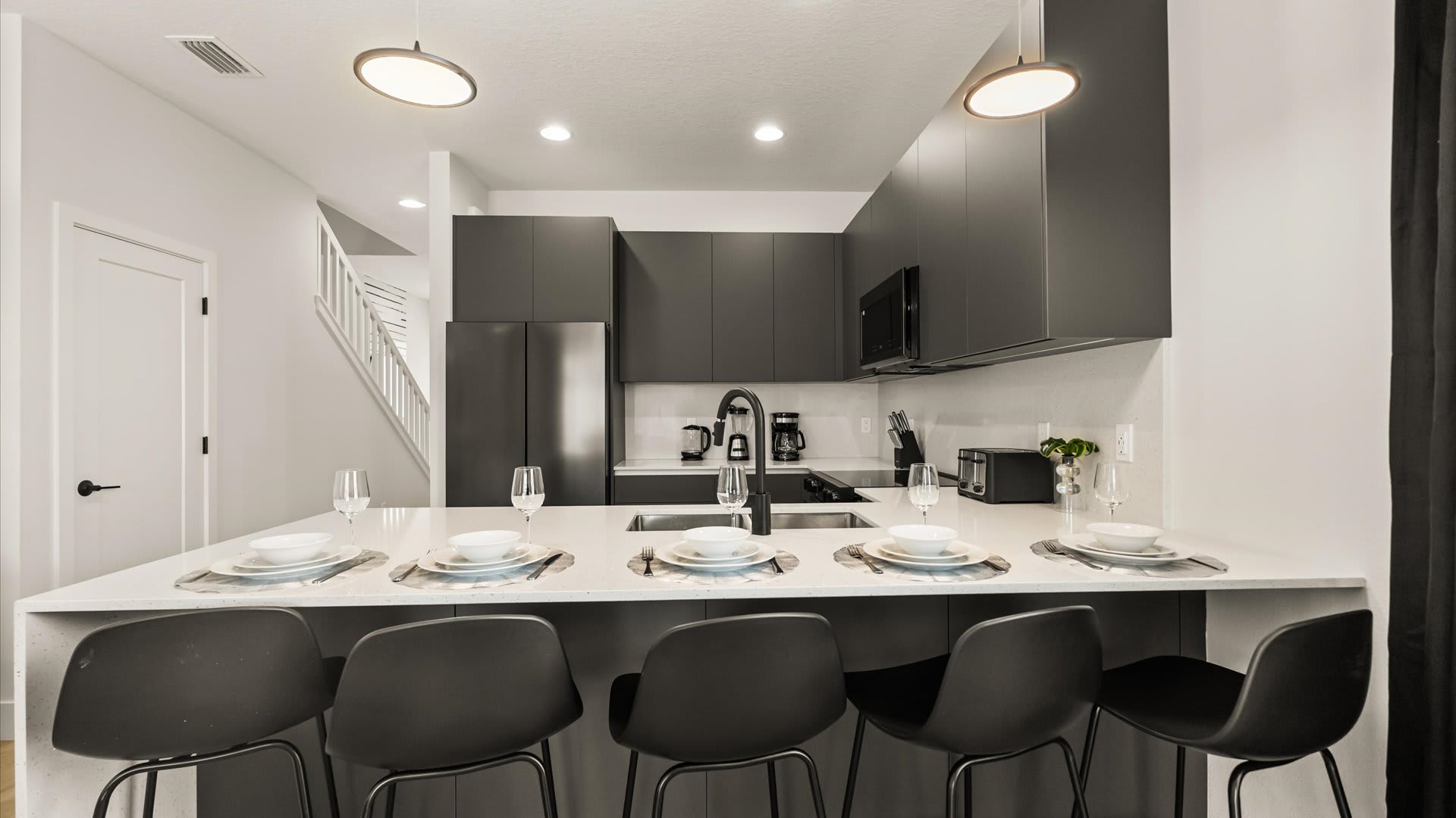 Wide shot of the kitchen island with four bar stools, set with place settings, overlooking the modern kitchen with dark gray cabinetry and stainless steel appliances, and a view towards a staircase.