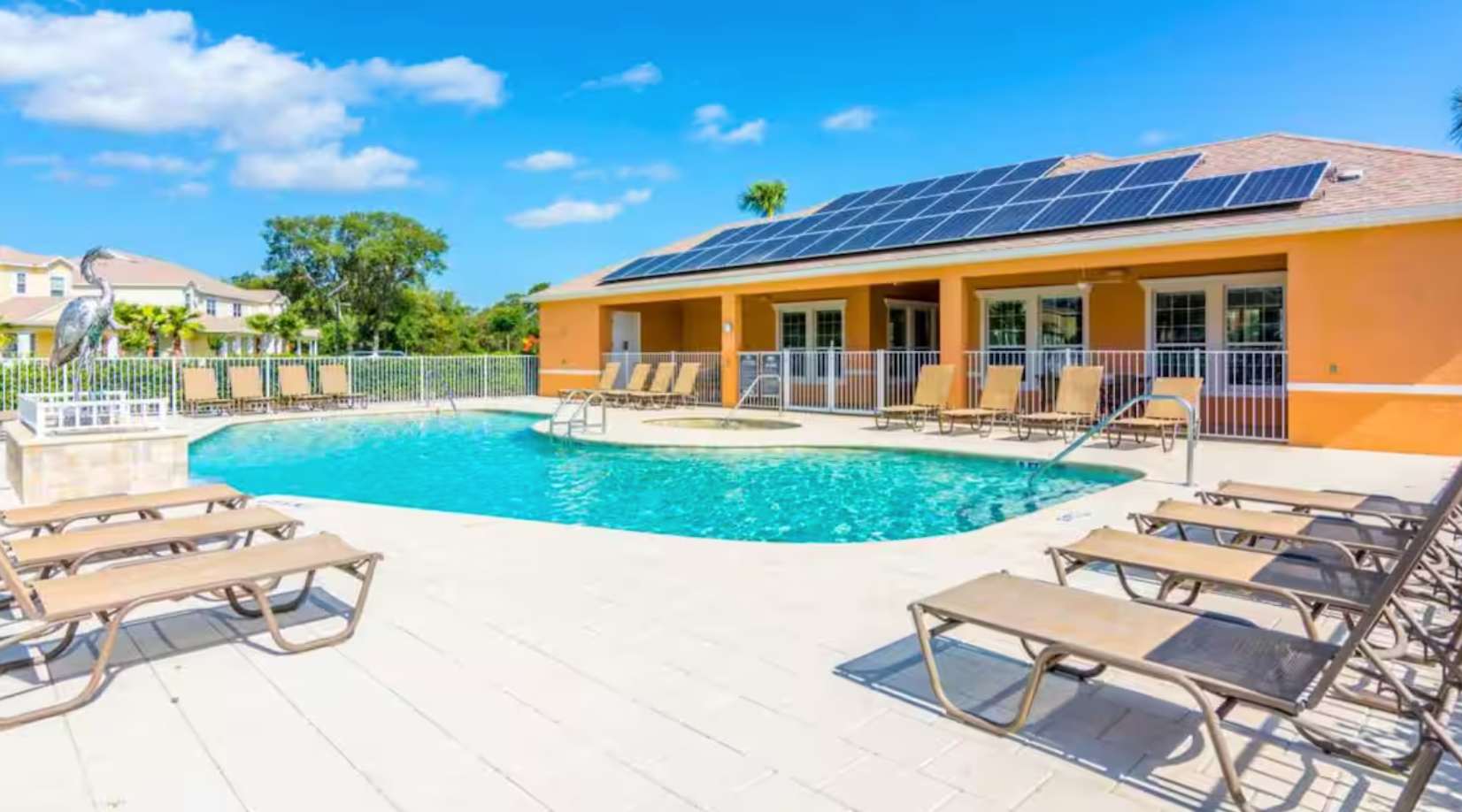 A spacious outdoor pool area featuring a large swimming pool, an integrated hot tub, and numerous lounge chairs on a light paver deck, with a clubhouse-style building topped with solar panels in the background.