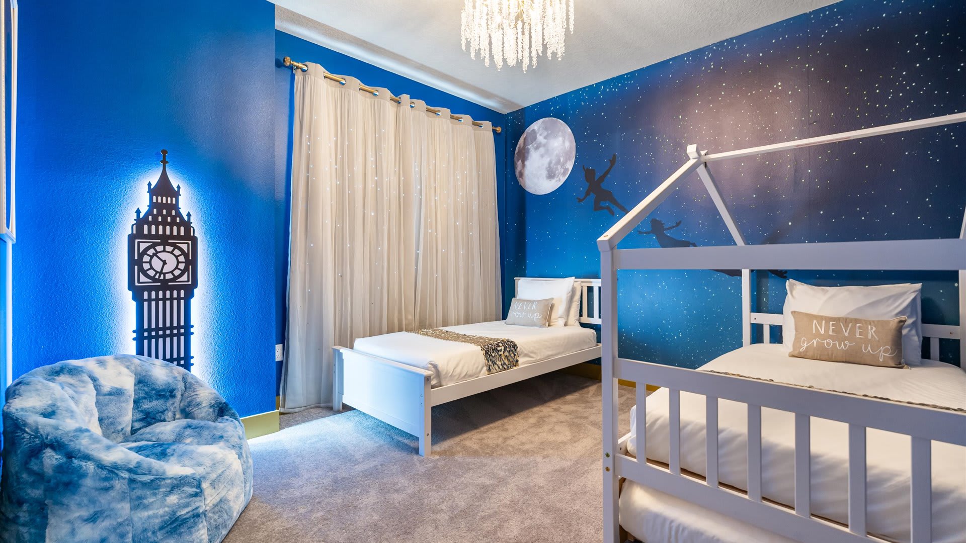 Peter Pan themed bedroom with deep blue walls featuring a starry night mural and a large moon. Two white twin beds, one with a house-frame, and a Big Ben-shaped light fixture create a magical atmosphere.