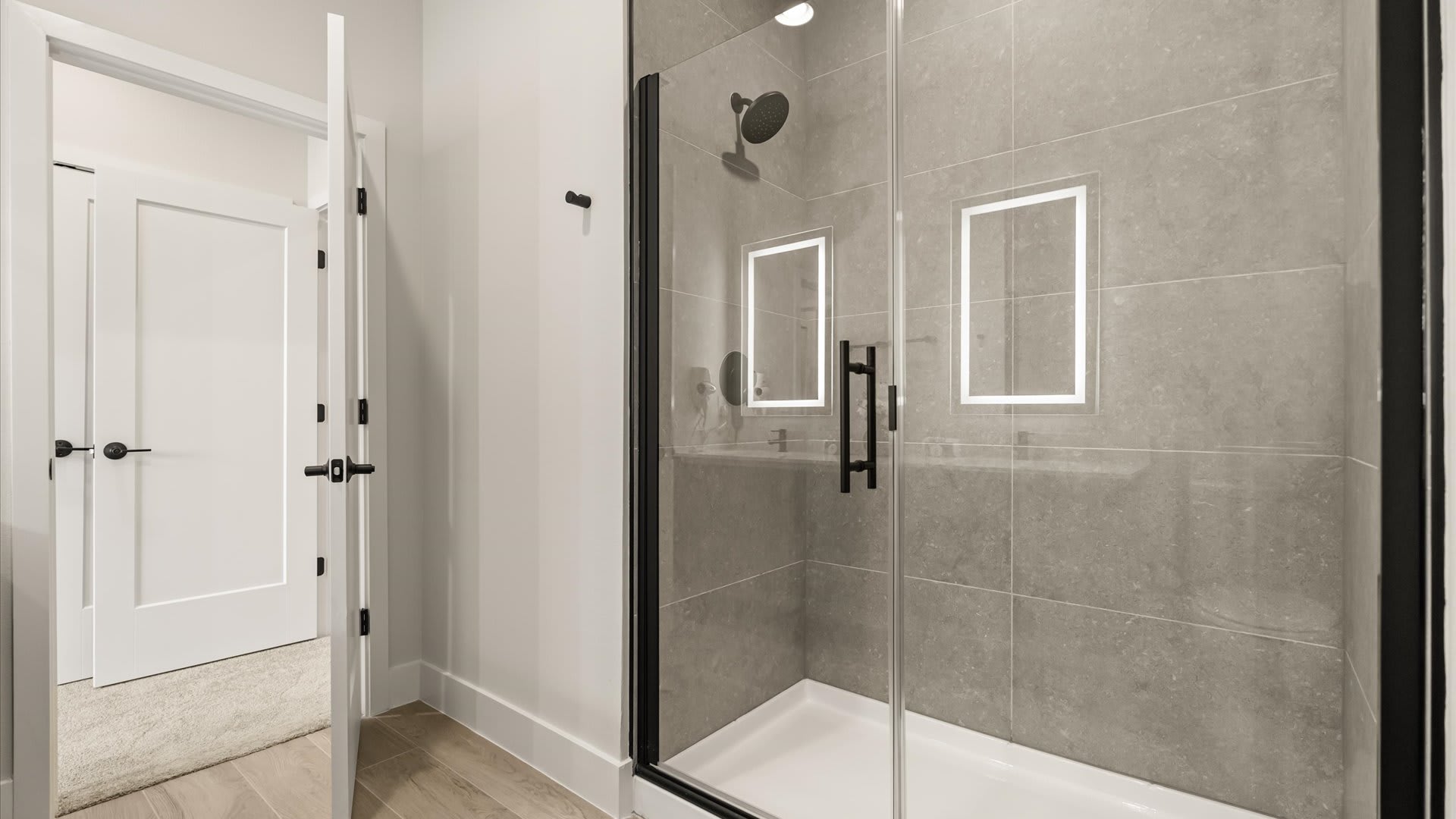 Contemporary walk-in shower with gray tiled walls, a glass enclosure with black framing, and a rain showerhead, featuring two illuminated niches and light wood-look flooring.