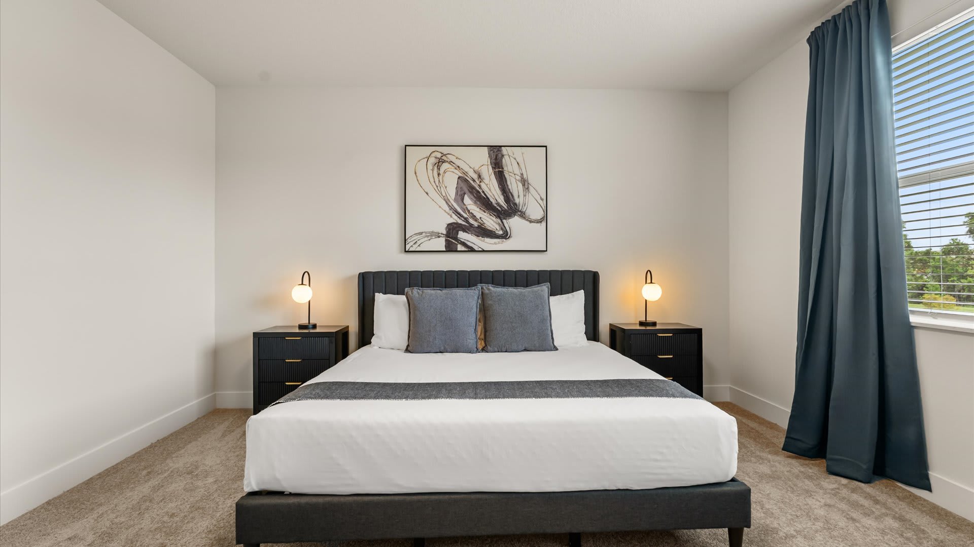 Serene bedroom with a dark gray upholstered bed, two black nightstands with modern lamps, and abstract artwork above the headboard, featuring light carpet and blue curtains.