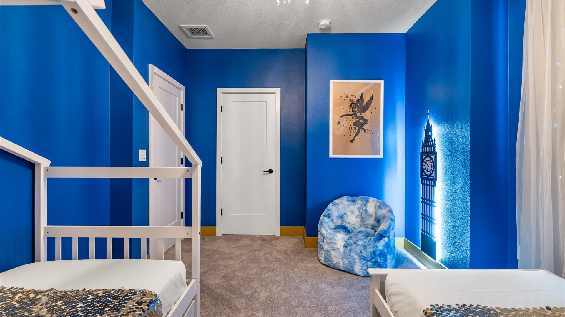 Another view of the Peter Pan themed bedroom, showcasing the vibrant blue walls with a starry mural and a Big Ben light fixture. A white house-frame twin bed and a blue tie-dye bean bag chair are visible.