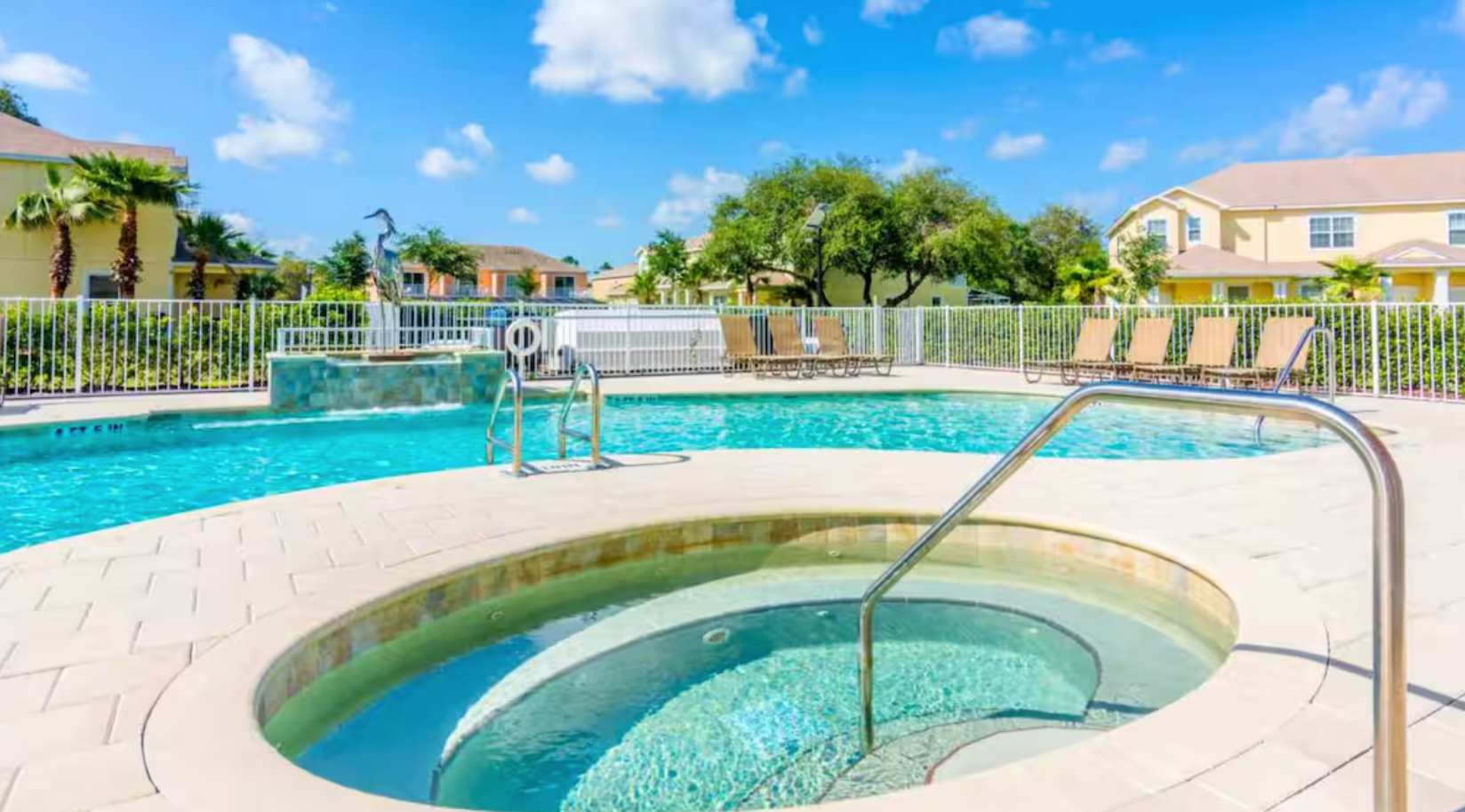A close-up view of a circular hot tub with light blue tiled steps, integrated into a light-colored paver deck, adjacent to a larger swimming pool with a metal handrail for entry.