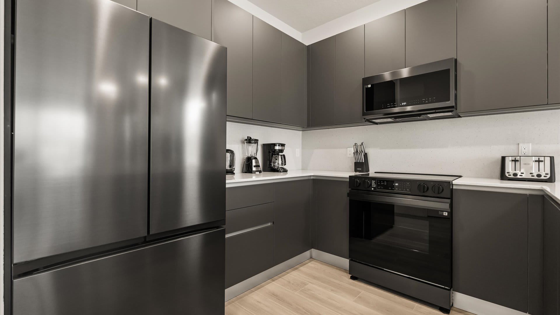 Corner view of the modern kitchen with dark gray flat-panel cabinets, white quartz countertops, and stainless steel appliances, including a large French door refrigerator, microwave, and electric range.