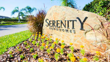 Large stone sign for "Serenity Townhomes" resort, surrounded by green foliage and yellow flowers under a bright sky. Palm trees are visible in the background.