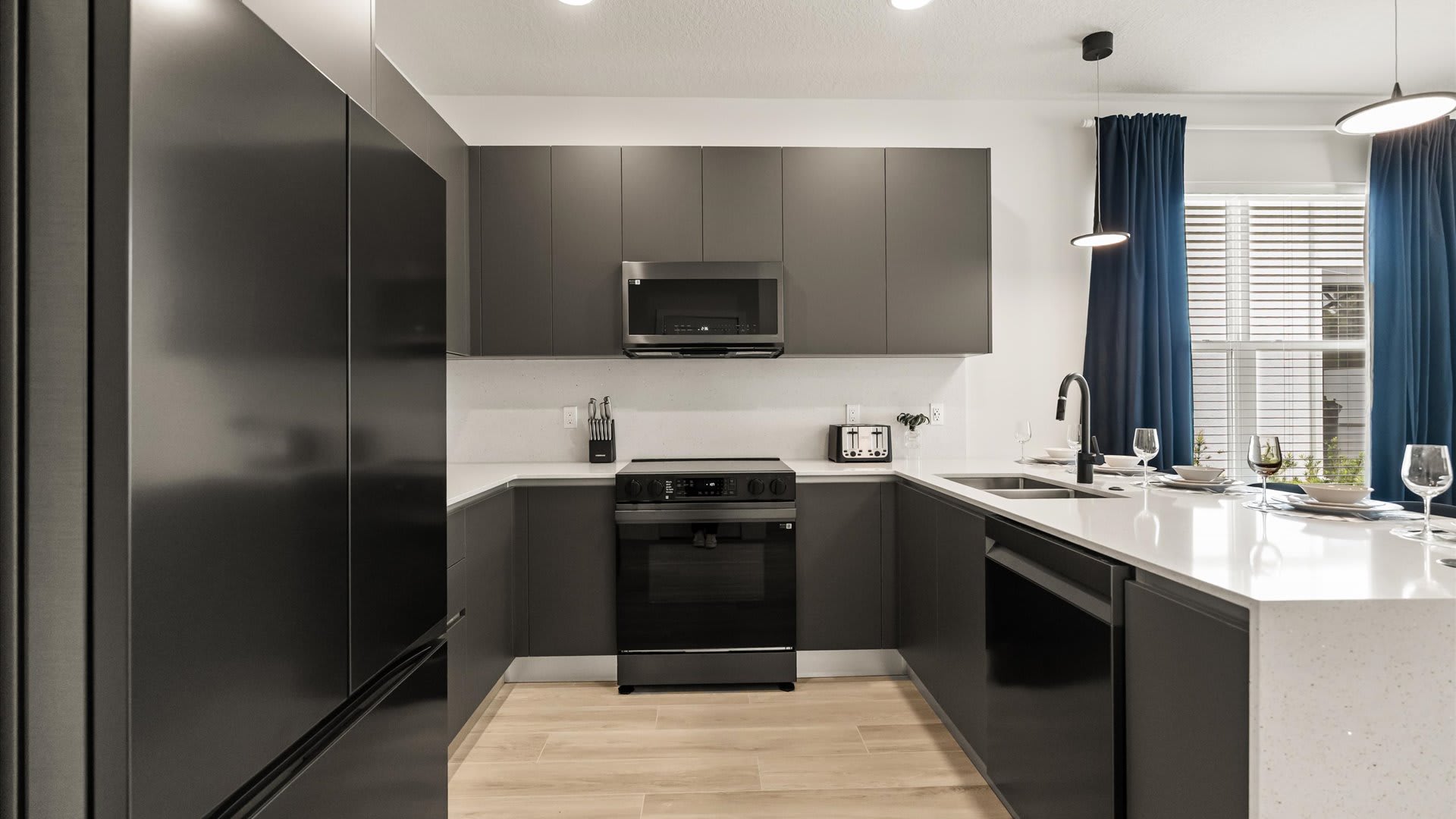Modern kitchen featuring dark gray flat-panel cabinetry, white quartz countertops, and stainless steel appliances, including a refrigerator, microwave, and electric range, with light wood-look flooring.