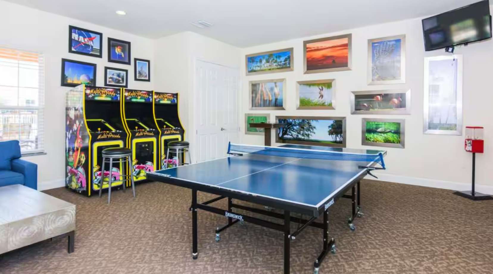 A vibrant game room featuring a blue ping pong table in the center, flanked by two classic arcade machines with stools, and walls adorned with various framed artwork and photos.