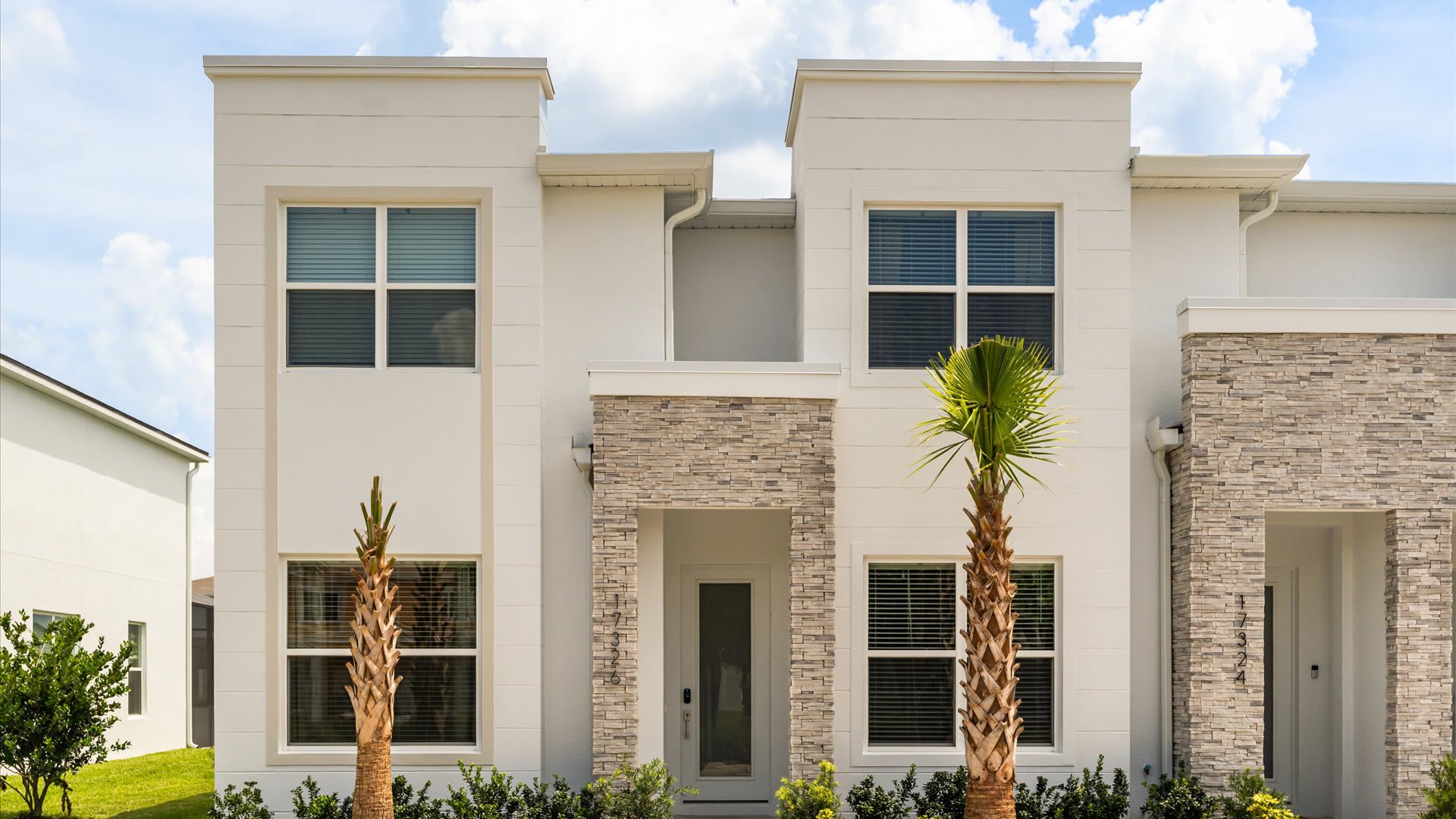Modern white townhouse exterior with stone accents around the entrance and lower windows. Two tall palm trees flank the front door, set against a clear blue sky with scattered clouds.