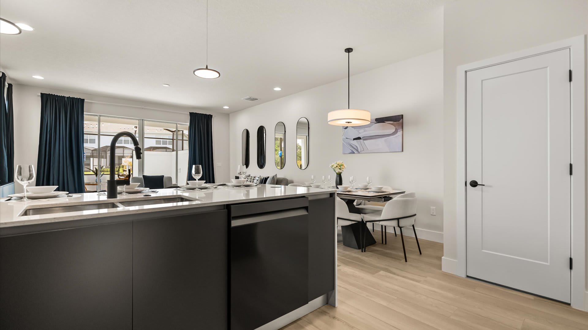 Open-concept view of the kitchen island with seating, leading into a dining area with a modern table and chairs, and a living space beyond, all featuring light walls and wood-look flooring.