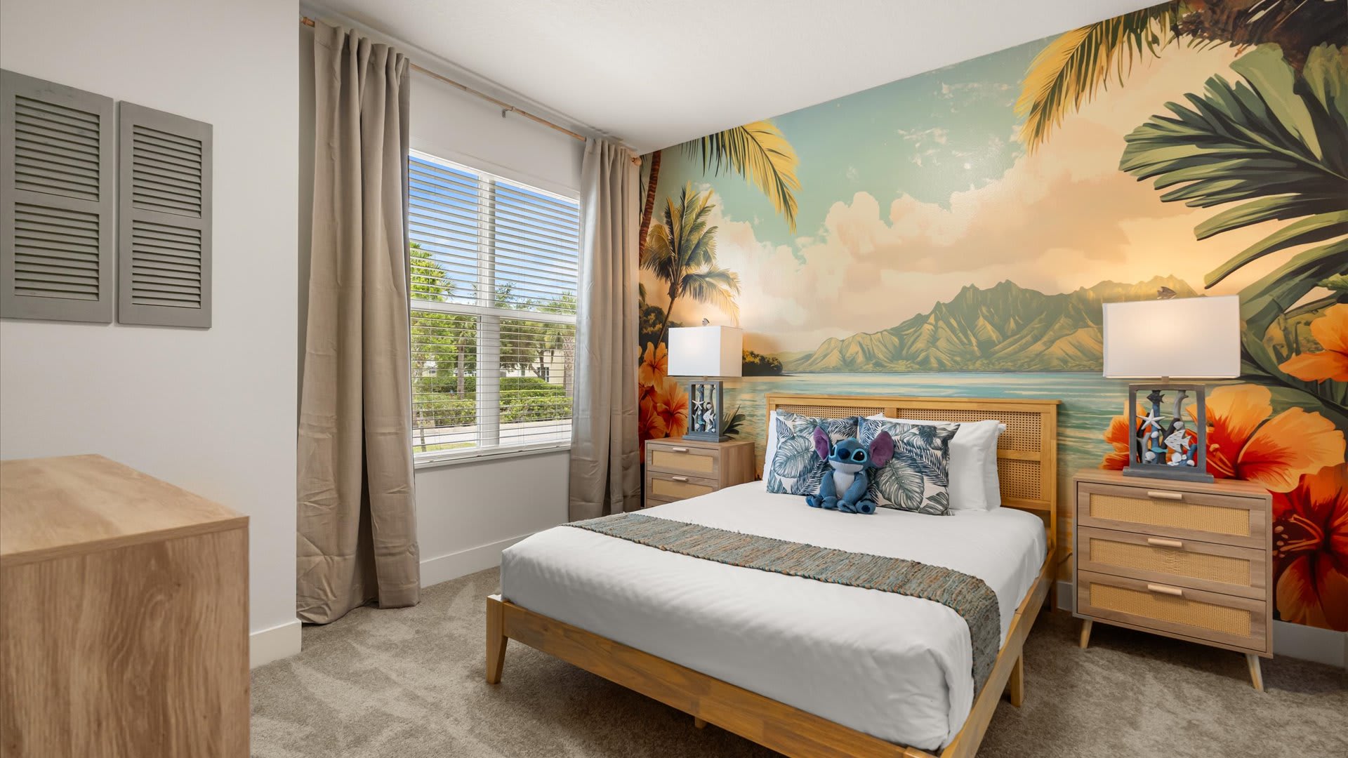 Themed bedroom with a vibrant tropical mural covering one wall, a light wood bed with cane accents, two matching nightstands with lamps, and light carpet flooring.