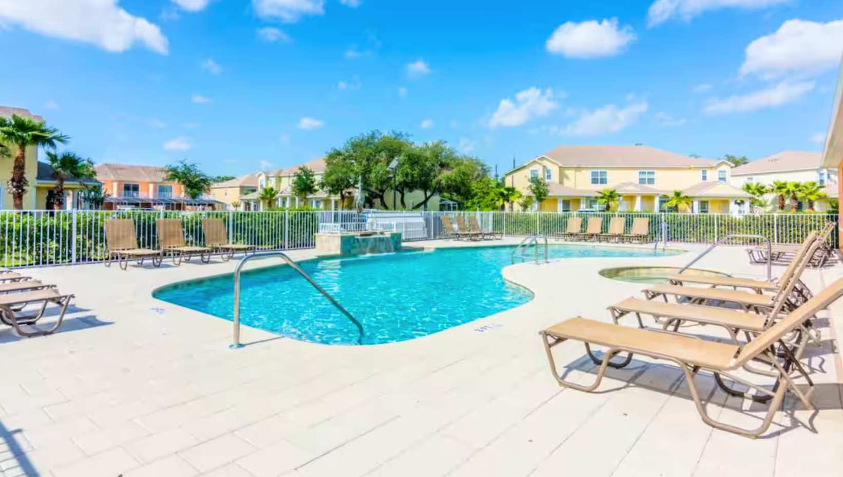 An inviting outdoor swimming pool with a curved shape, surrounded by a light-colored paver deck and multiple brown lounge chairs, enclosed by a white metal fence with residential buildings and trees in the distance.