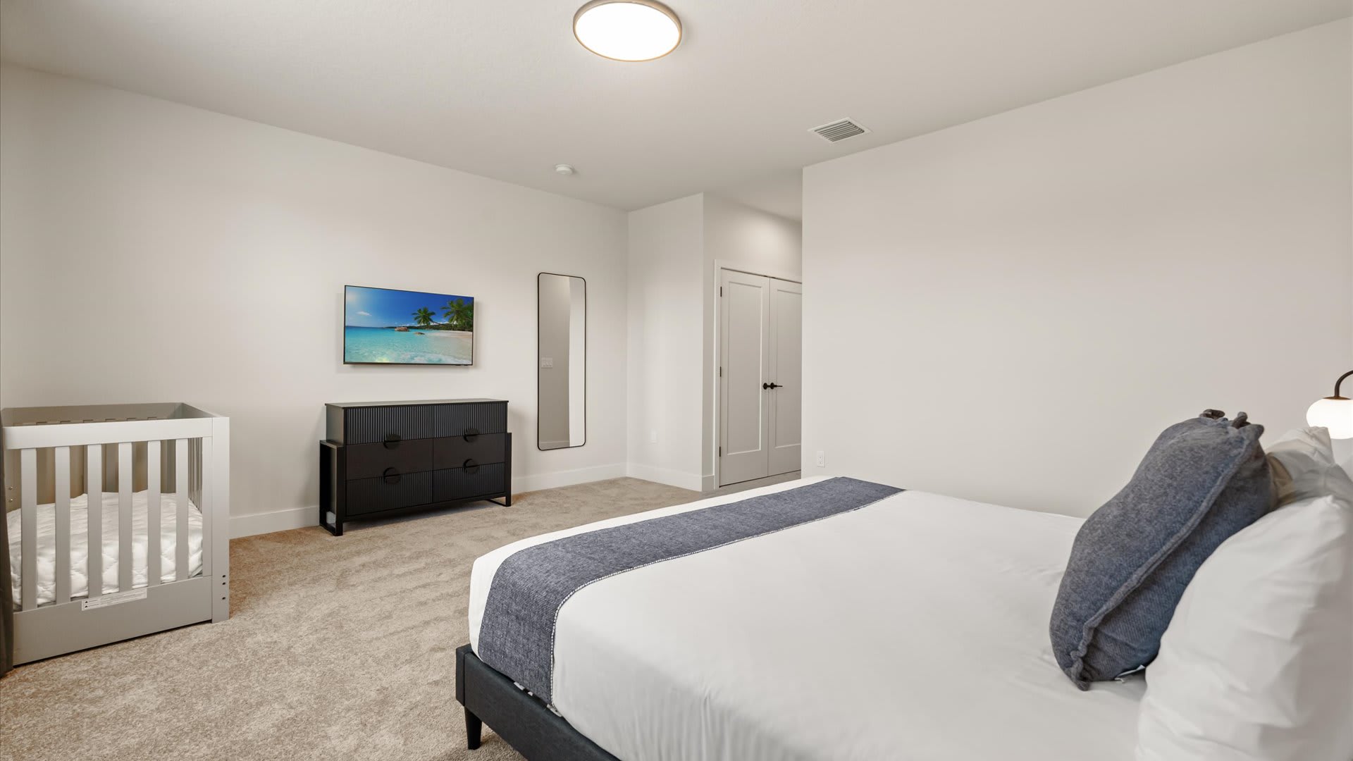 View of the bedroom showing a crib, a black dresser with a TV mounted above it, and a full-length mirror, with light carpet and a white door leading to a closet.