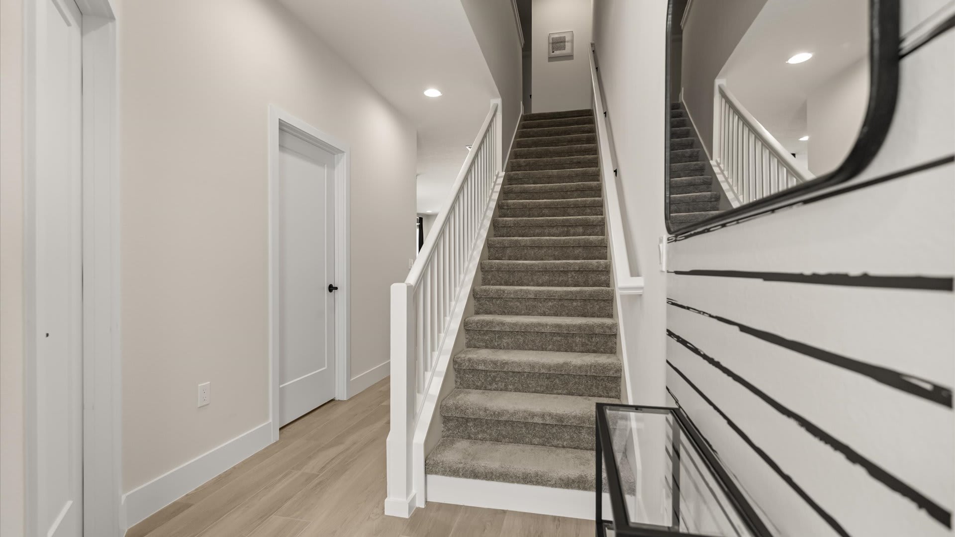 Bright entryway featuring a carpeted staircase with white railings, light wood-look flooring, and a decorative console table with a large oval mirror, leading to a white door.