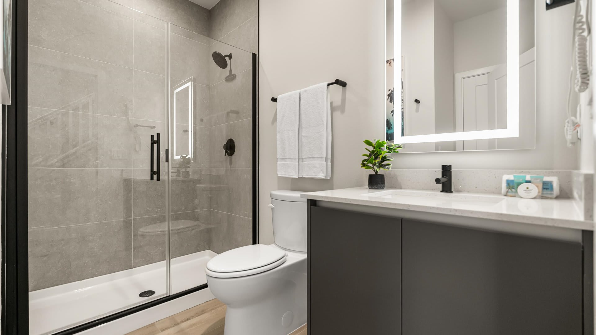 Modern bathroom featuring a glass-enclosed walk-in shower with grey tiled walls and a black shower head. A vanity with a white countertop, dark grey cabinet, and an illuminated mirror is visible.
