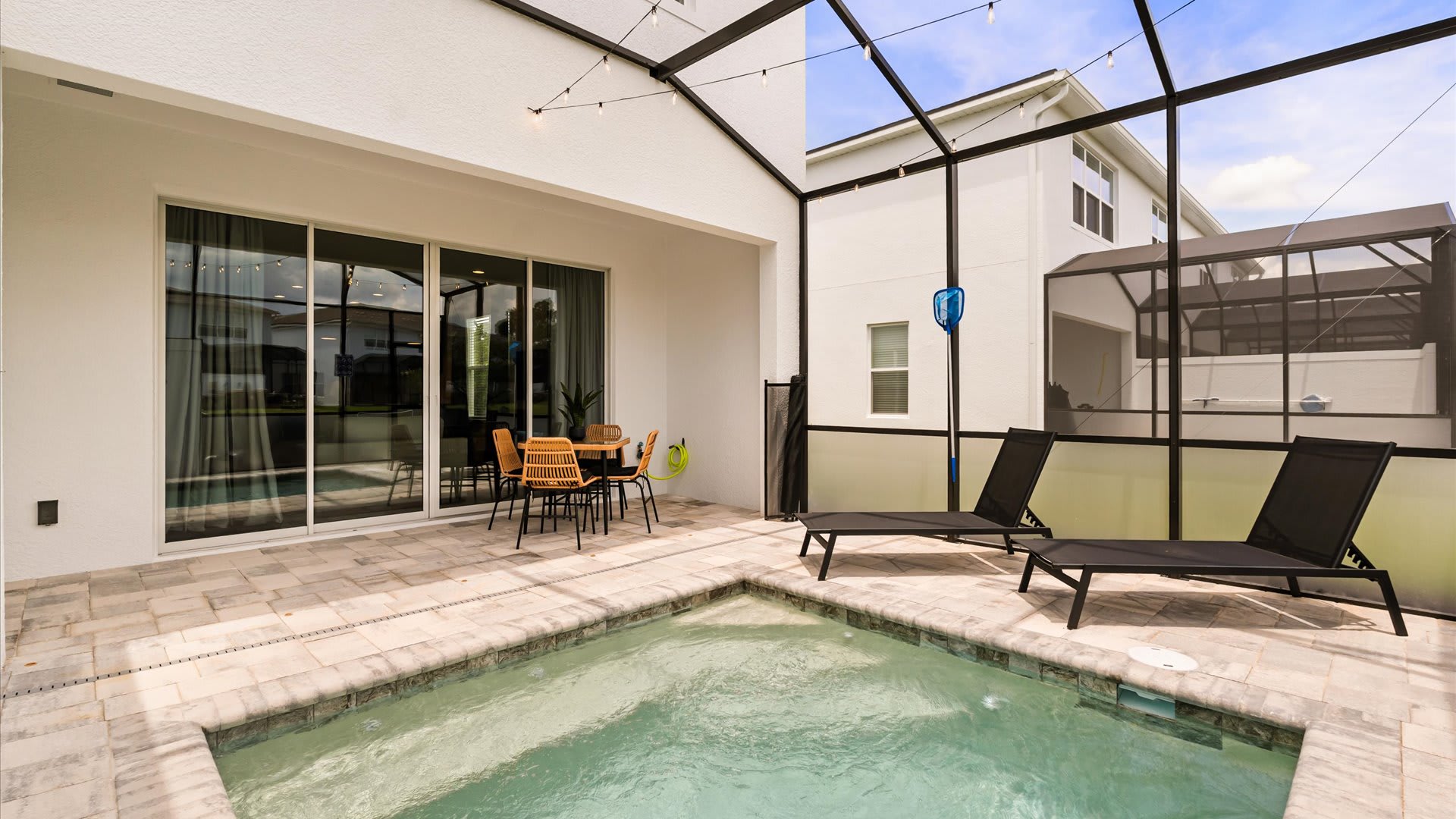 Wide view of the screened-in patio with a small pool, two black lounge chairs, and an outdoor dining table, showing the surrounding residential area through the screen.