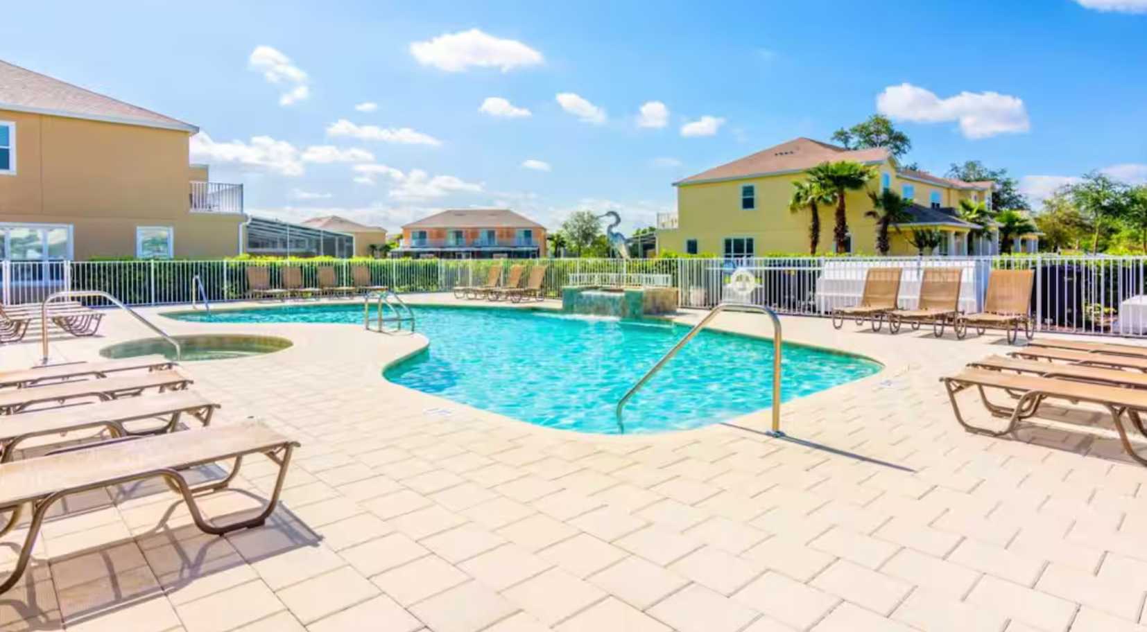 A bright outdoor swimming pool with light blue water, surrounded by light-colored paver decking and numerous brown lounge chairs, set against a backdrop of yellow and beige residential buildings under a clear blue sky.